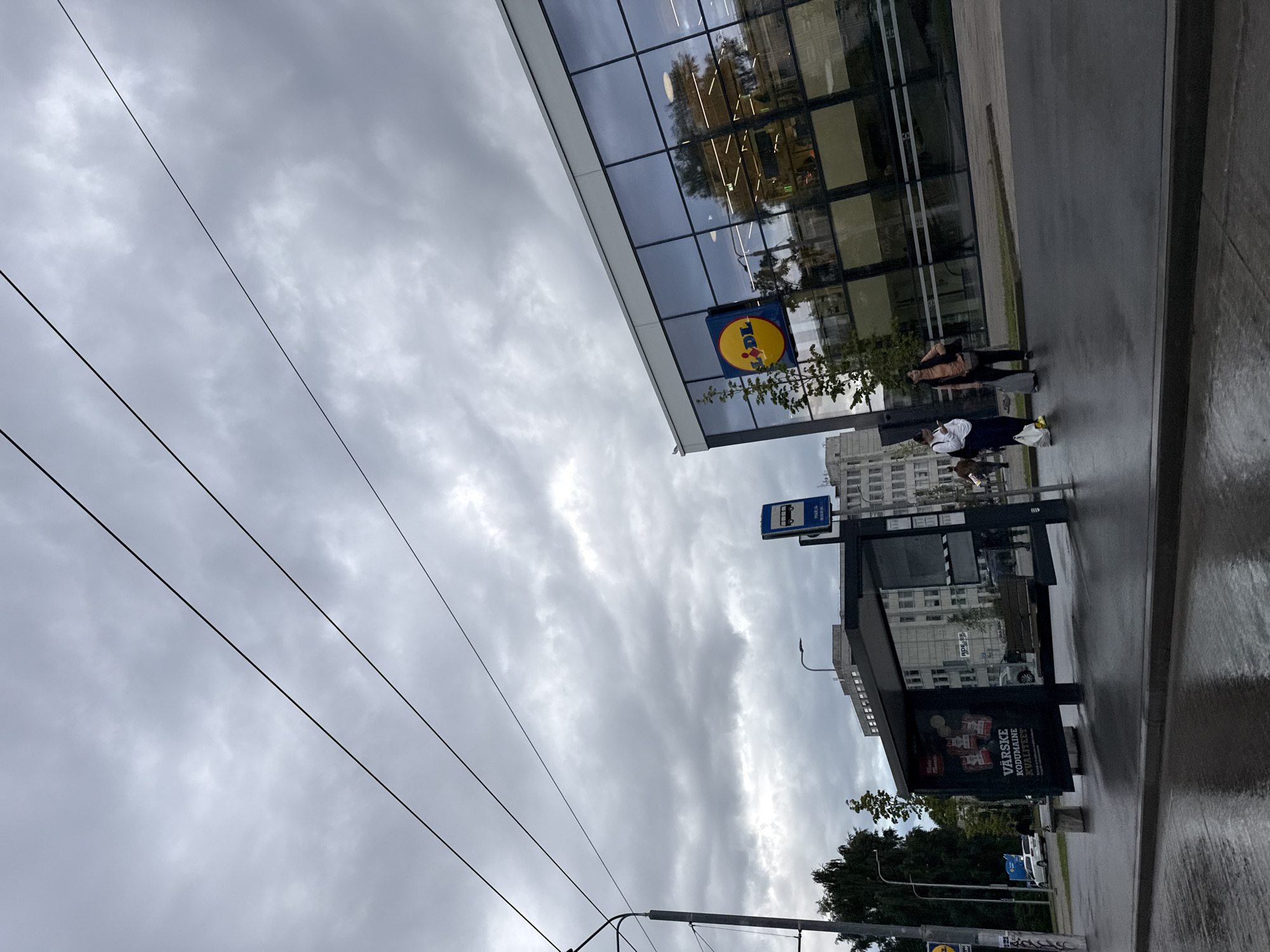 The image depicts an overcast day in Tallinn, Estonia, showing a street scene with a bus stop and a Lidl supermarket. The perspective is from street level, looking slightly upwards and to the right. In the foreground, the wet asphalt road reflects the cloudy sky. To the left, a modern bus stop shelter with advertising posters is visible, one of which reads "VÄRSKE KODUMAISE KVALITEET". A lamppost is situated behind the bus stop. Further back, tall buildings are visible, and trees line the street on the left. On the right side of the image, a glass-fronted Lidl supermarket with its distinctive yellow and red logo is prominent. Two individuals, a woman in a white top and dark trousers with a shopping bag, and another woman in an orange top and dark vest with a shopping bag, are standing on the pavement near the supermarket entrance. Overhead, thick, dark clouds fill the sky, with power lines crisscrossing the frame.