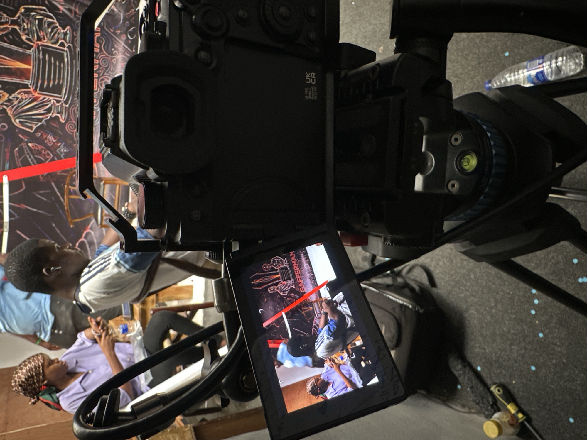 A high-angle view focuses on a large black digital camera, likely a cinema camera, mounted on a tripod, set up to film two individuals seated in what appears to be a small interior space. The camera's display screen reflects the scene it's capturing: two individuals, a woman wearing a headscarf and a light purple shirt, and a man in a blue and white shirt, seated next to each other. A water bottle is visible on the table they are at. A black bag rests on the floor near the tripod legs, along with a small yellow container and a tool. A red tape or marking is seen on the wall in the background, which features a mural or drawing. The location is within Ikeja, Nigeria.
