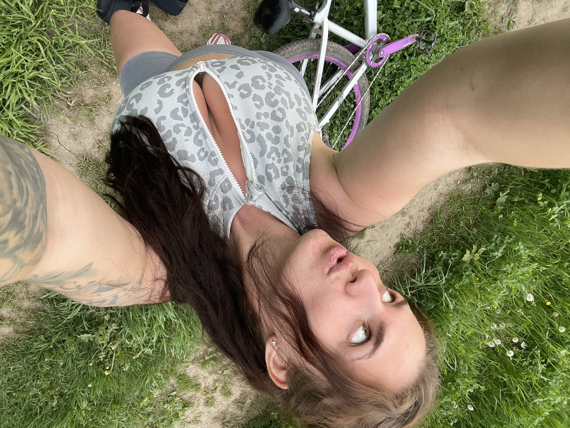 A woman is lying on her back in a grassy area outdoors. She is wearing a gray and white patterned top with a zipper that is partially open, revealing some cleavage. She has gray shorts on as well. A white bicycle with a purple chainring is visible near her. There is a tattoo on her left arm. The location cannot be confirmed as Ostrov, Russia, based on the provided image alone.