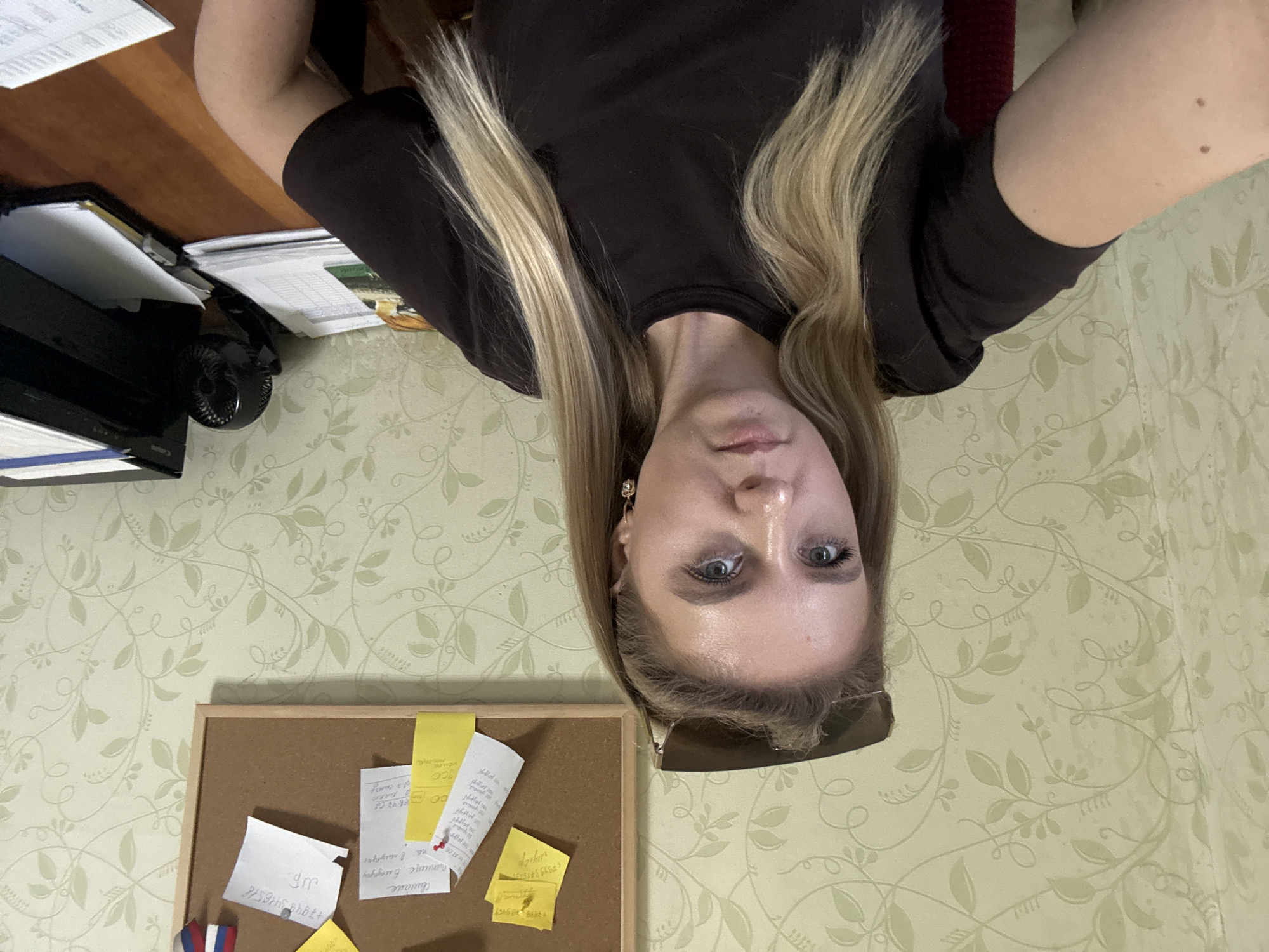 In an indoor setting with floral wallpaper, a young woman with blonde hair sits at a desk. She is wearing a dark t-shirt and has sunglasses perched on her head. She is looking directly at the camera. To her right, on a corkboard, are several sticky notes and a small Russian flag. A printer and a stack of papers are visible on the desk. The scene appears to be a home office or workspace.