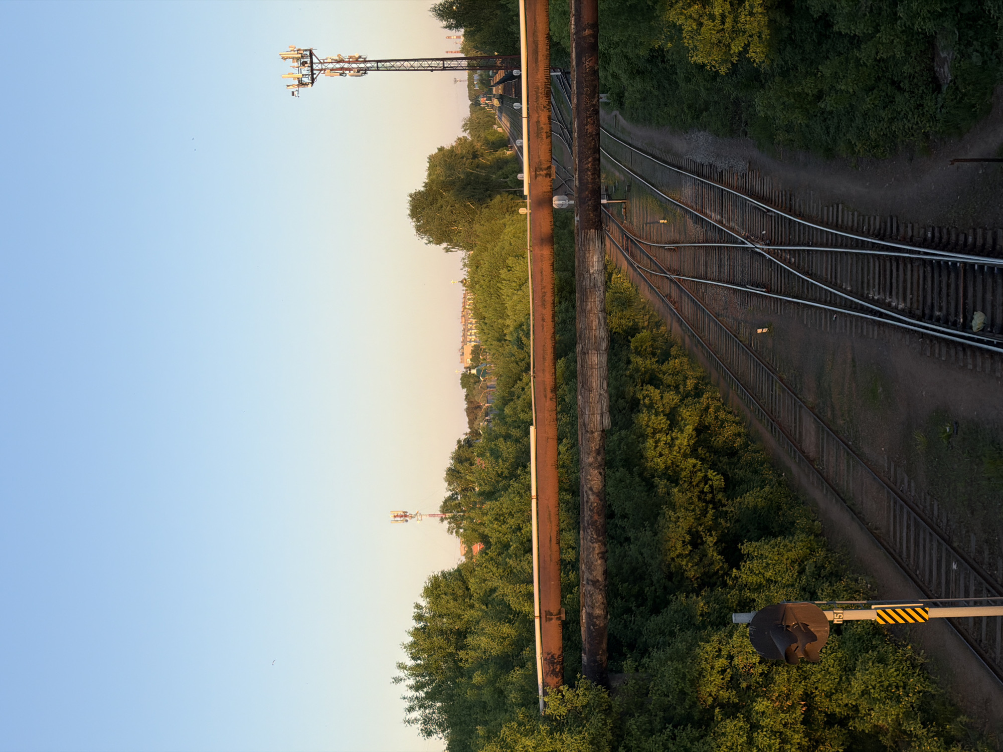 The image shows a view of railway tracks near Chaikovsky, Russia, likely taken from an elevated position. The tracks are multi-directional, with junctions visible amidst surrounding vegetation. Two metal pipes run vertically along the image's central portion. In the background, there are buildings and a communication tower. A railway signal is visible at the lower right. The sky is light blue.