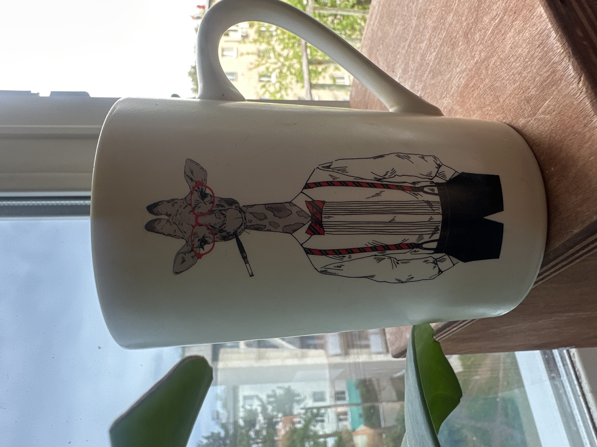 A mug with a giraffe wearing glasses and a suit is on a wooden surface near a window. Outside the window, buildings and trees are visible, possibly indicating a location in the city of Chaikovsky, Russia. A green plant is partially visible at the bottom of the frame.