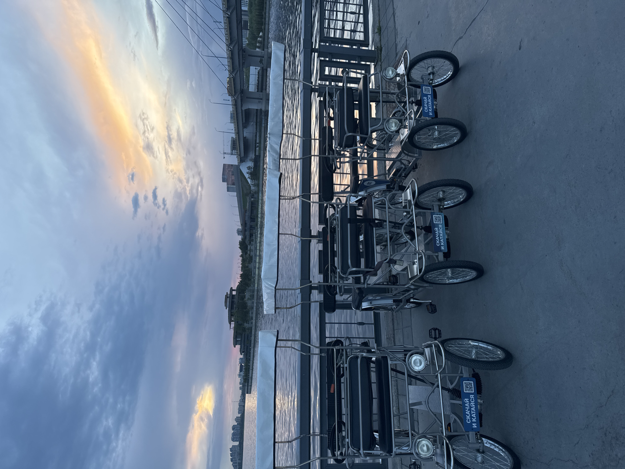 The image shows a scene at a waterfront location in Kazan, Russia. There are three quadricycles (four-wheeled bicycles) parked on a paved surface next to a metal railing. The railing separates the pavement from the water. In the background, there is a bridge and buildings, indicating an urban setting. The sky is visible, with colors suggesting either sunrise or sunset. The quadricycles have signs with Russian text and QR codes. The lighting suggests it is either early morning or late evening. No people are visible in the frame.