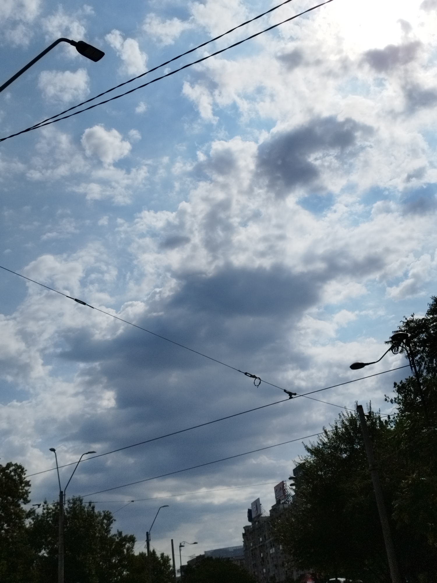 The image captures a street scene in Bucharest, Romania, under a cloudy sky. The perspective is looking upwards, dominated by the sky and overhead wires. Several streetlights are visible, some silhouetted against the bright sky, and others partially obscured by the trees and buildings. The sky is a mix of blue patches and various shades of grey clouds, with the sun partially visible through the cloud cover, creating a bright area towards the top right. Overhead wires, likely for streetcars or power, crisscross the frame. In the lower portion of the image, the dark silhouettes of trees line the street, and the upper parts of buildings are visible, one with visible signage. The overall impression is an urban environment during the day, with a dramatic cloudy sky.