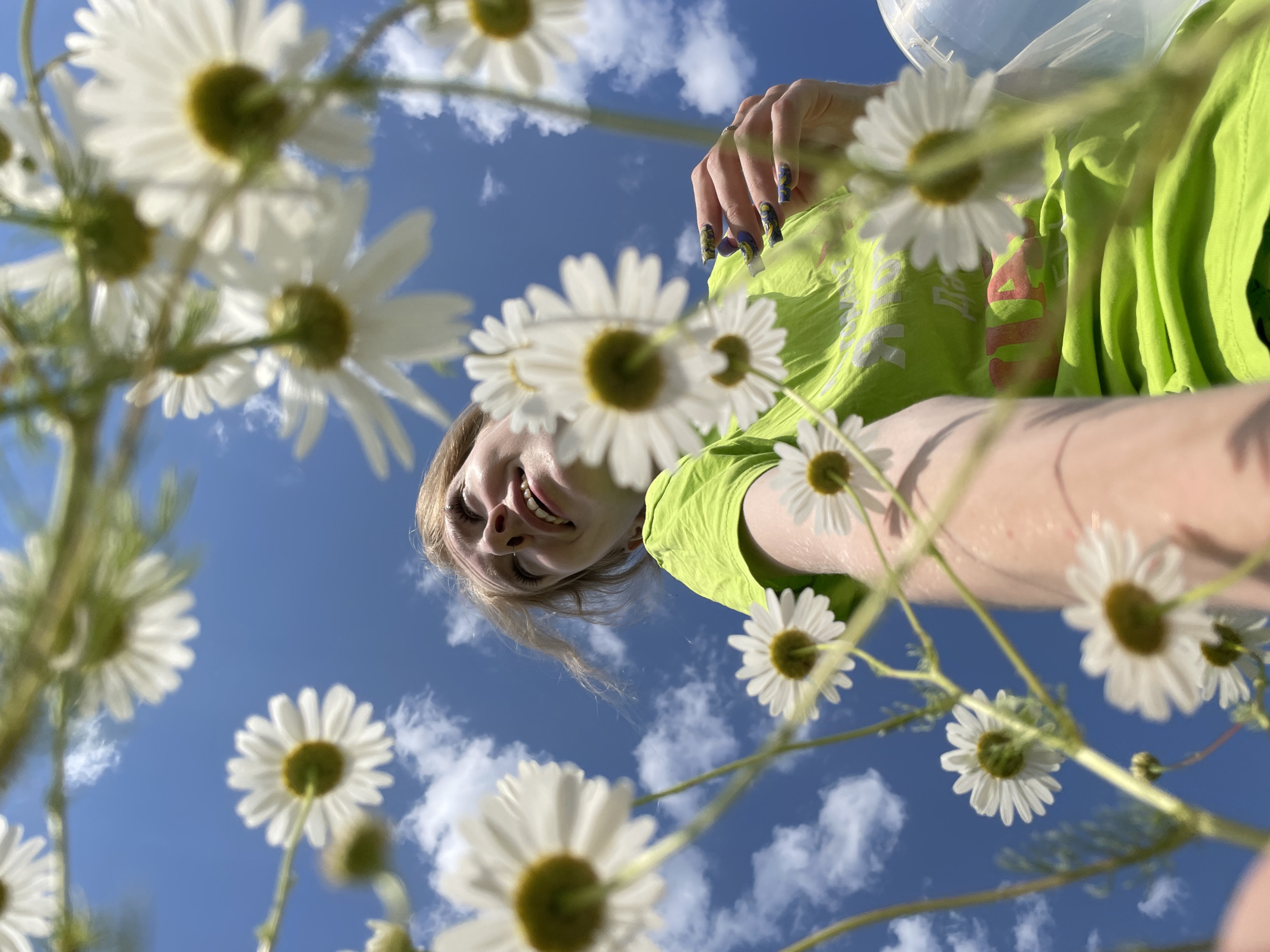 The image, taken from a low angle, features a young woman with blonde hair looking up at the sky, her mouth open in a smile. She is wearing a bright green t-shirt. The foreground is filled with white daisies with yellow centers, some in focus and some blurred, creating a natural frame. The background is a bright blue sky with scattered white clouds. The perspective suggests the photograph was taken outdoors, possibly in a field or park, with the camera positioned beneath the daisies, looking upwards towards the woman and the sky. The overall impression is one of a sunny, cheerful day. The provided location is Сям-Можга, Russia.