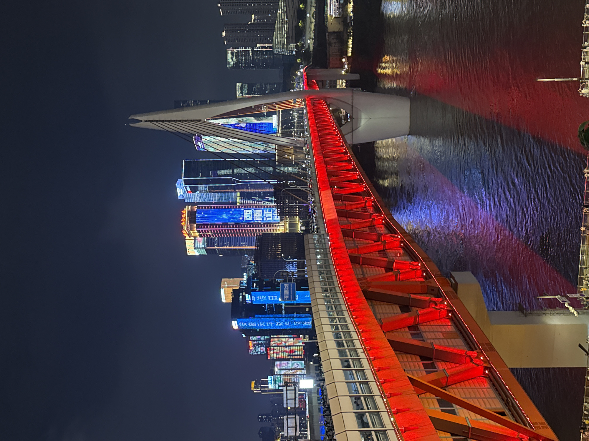 This is a nighttime, eye-level shot of the Hongyadong Bridge, also known as the Qibao Bridge, in Yuzhong District, China. The bridge, a suspension bridge with a distinctive white pylon, is illuminated with bright red lights along its main structure and deck. The deck of the bridge has a walkway with railings, and there are numerous people visible on the walkway, suggesting it is a popular pedestrian area. The water below the bridge reflects the vibrant red lights of the bridge and the colorful lights of the surrounding cityscape. In the background, several modern skyscrapers are lit up with a variety of colors, including blues, oranges, and purples, showcasing a dense urban environment. The sky is dark, indicating it is nighttime.