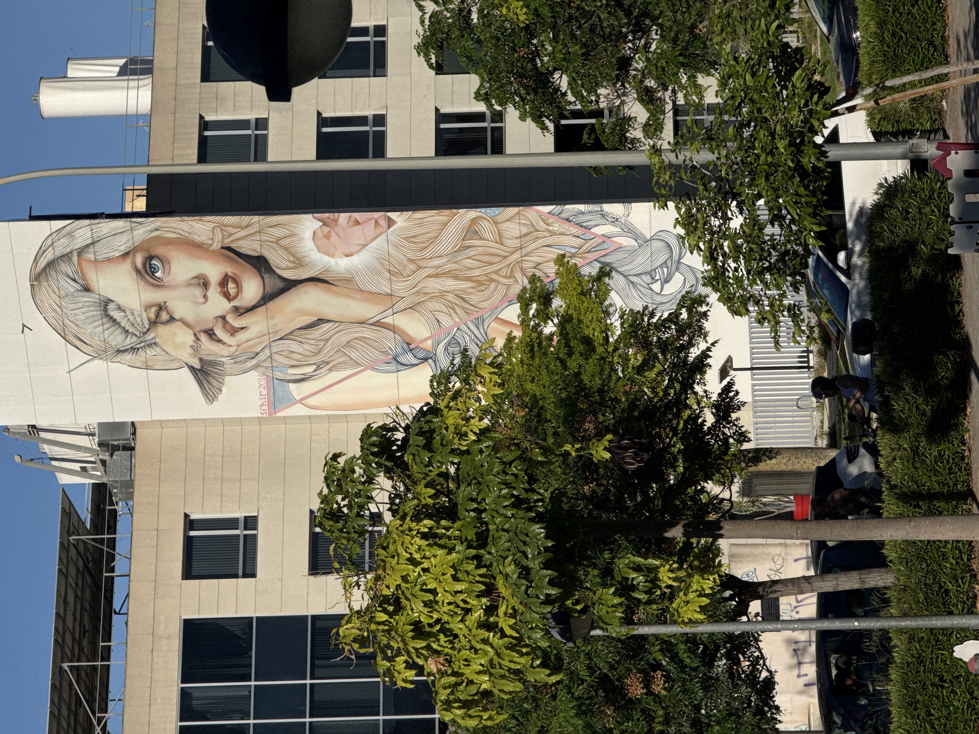 A large mural adorns the side of a building, depicting a woman with flowing, light-colored hair. A bird-like creature is near her face, and a geometric shape is above her head. The building has a beige facade with dark-framed windows. Green trees partially obscure the view of the mural. A streetlight and parked cars are visible to the right of the building. A person sits near the cars. The image is taken from ground level. The building's location is Unknown, Unknown.