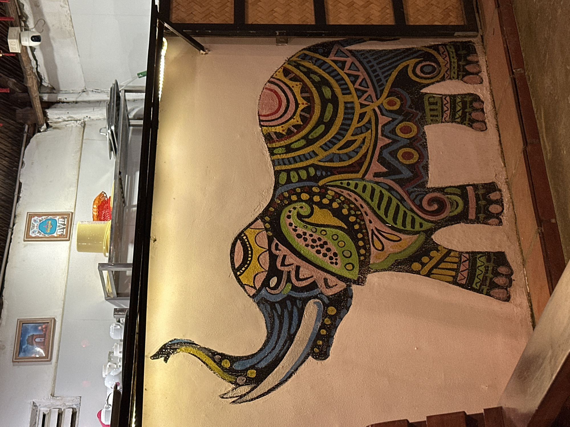 The image depicts the interior of what appears to be a cafe, restaurant, or shop, characterized by a vibrant and artistic aesthetic. The main focal point is a large, highly stylized mural of an elephant painted on a light-colored wall. The elephant is depicted in profile, facing left, and is adorned with intricate geometric and organic patterns in a rich color palette of yellow, blue, green, pink, black, and red. A strip of artificial light runs vertically alongside the mural, illuminating it with a warm glow.
To the left of the mural, the room features other elements. A security camera is mounted high on the wall, near exposed wooden beams that suggest a rustic architectural style. Further down on the left wall, two framed pictures are visible. The upper frame contains a blue and white image with the word "WELCOME" clearly visible. Below it is another framed picture, though its details are less distinct.
Below these framed pictures, shelves hold various items, including several white cups and a yellow bucket or container. The ceiling area, or possibly an upper wall section, incorporates a woven material, likely bamboo or rattan, adding to the natural or traditional decor. The overall lighting suggests an indoor setting, possibly in the evening, relying primarily on artificial sources. No people are visible, and there is no active event taking place, presenting a static view of the decorated interior.