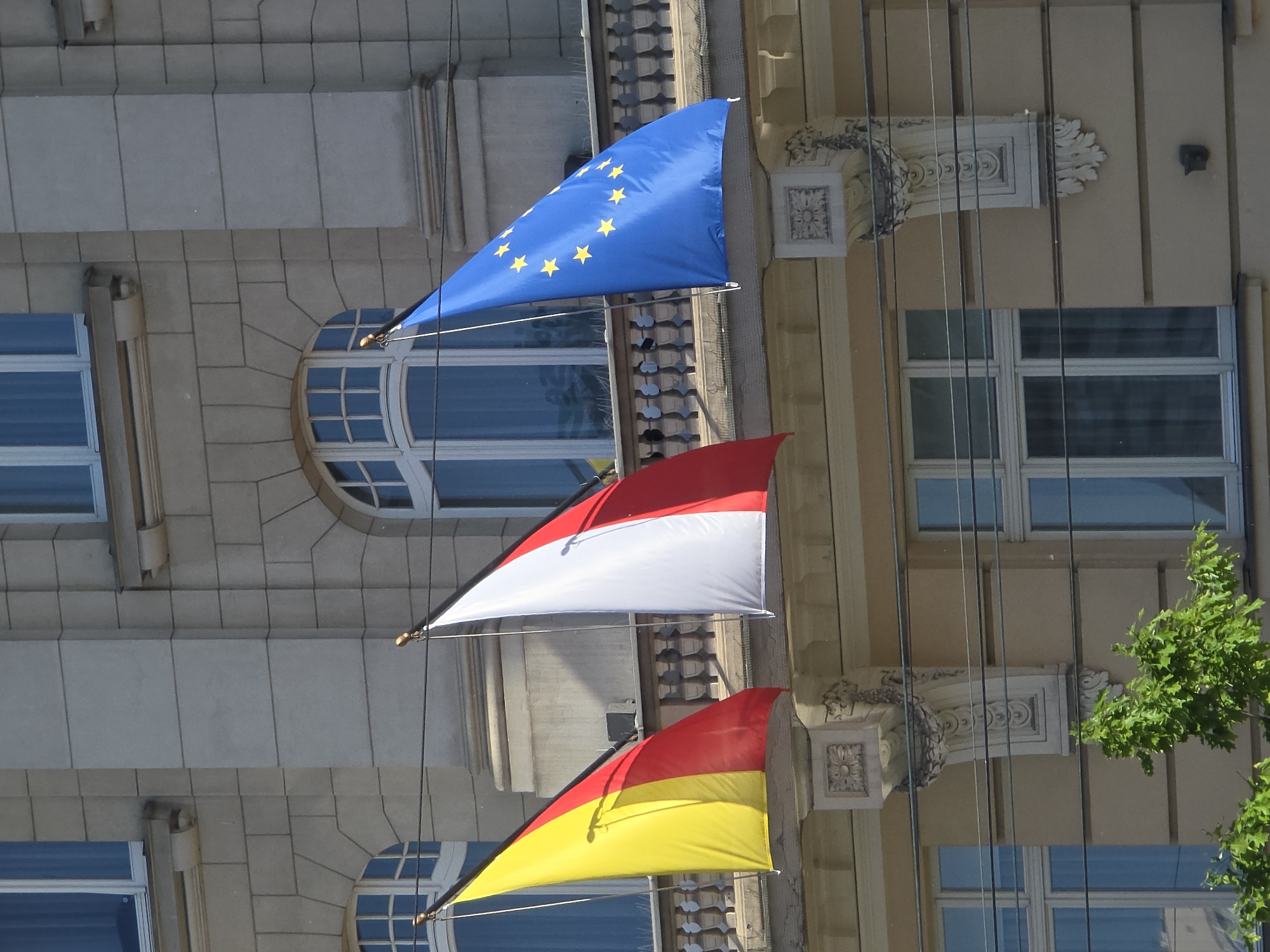 A vertical outdoor shot shows three flags of the European Union, Indonesia, and Germany hanging on a building in Warsaw, Poland. The blue EU flag with yellow stars is on top, followed by the red-and-white Indonesian flag, and the red-and-yellow German flag. The building has windows with arched tops and decorative elements on its facade. Wires run down the side of the building, partially obscuring the view. Green foliage is visible on the right side of the image.