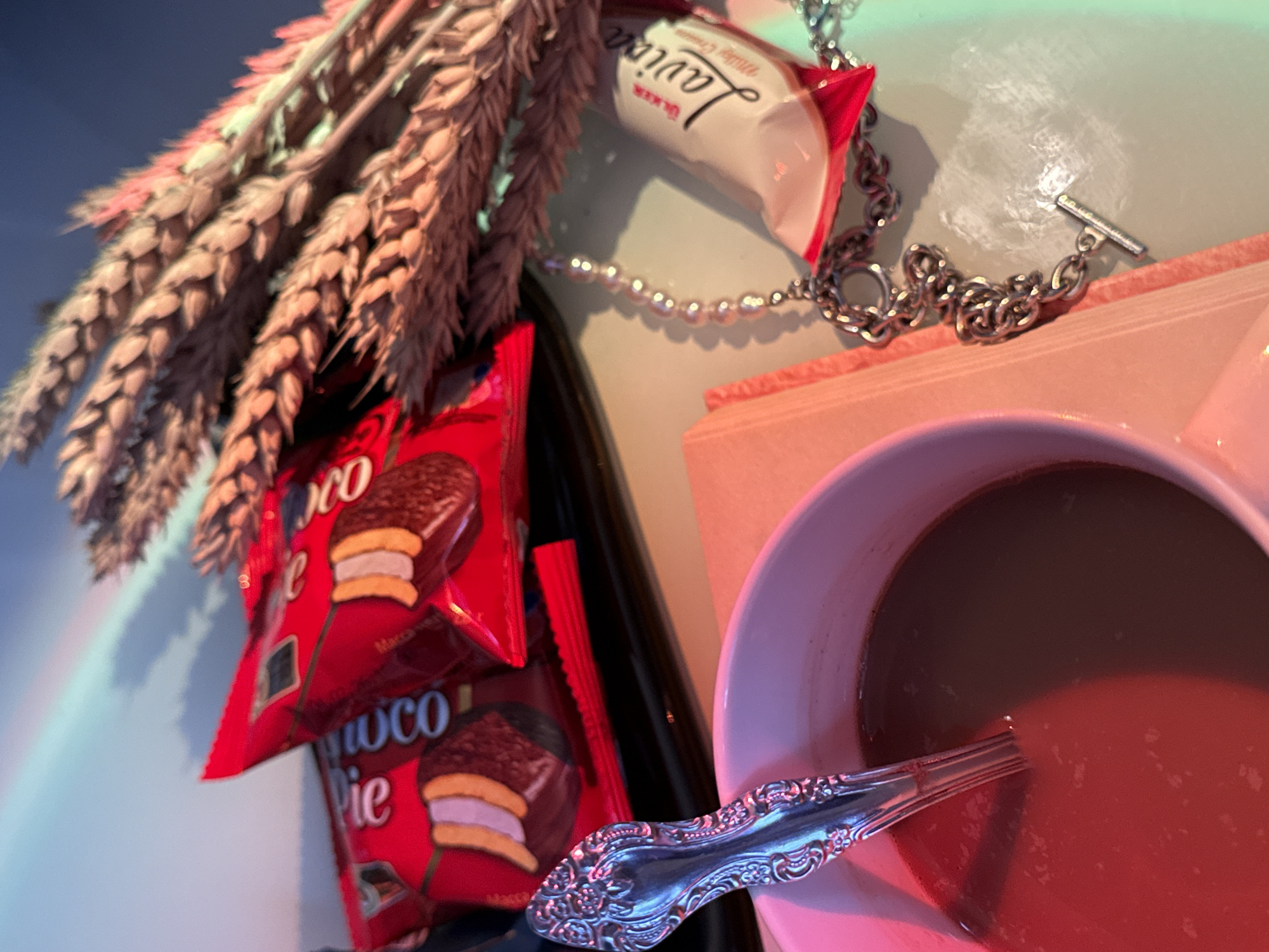 A tabletop still life is depicted, illuminated by multiple colored light sources. In the bottom-left, a light-colored mug contains a dark liquid with a silver-colored spoon featuring an ornate handle. Adjacent to the mug on the right, a light pink or beige book lies horizontally. Behind these items, two red packages of "Choco Pie" snacks are visible on a dark surface. To the right, a cluster of dried wheat stalks is positioned. A silver-toned necklace, composed of both a chunky chain and a segment of faux pearls with a T-bar clasp, is laid out on the surface. Next to the necklace, a white and red package of "ÜLKER Laviva Milky Cream" is present. The scene is cast with a reddish-pink light on the left and a greenish-blue light on the right. The upper background reveals a blurred blue sky with a distinct, multi-colored arc of light, resembling a rainbow.
Location: Unknown, Unknown
