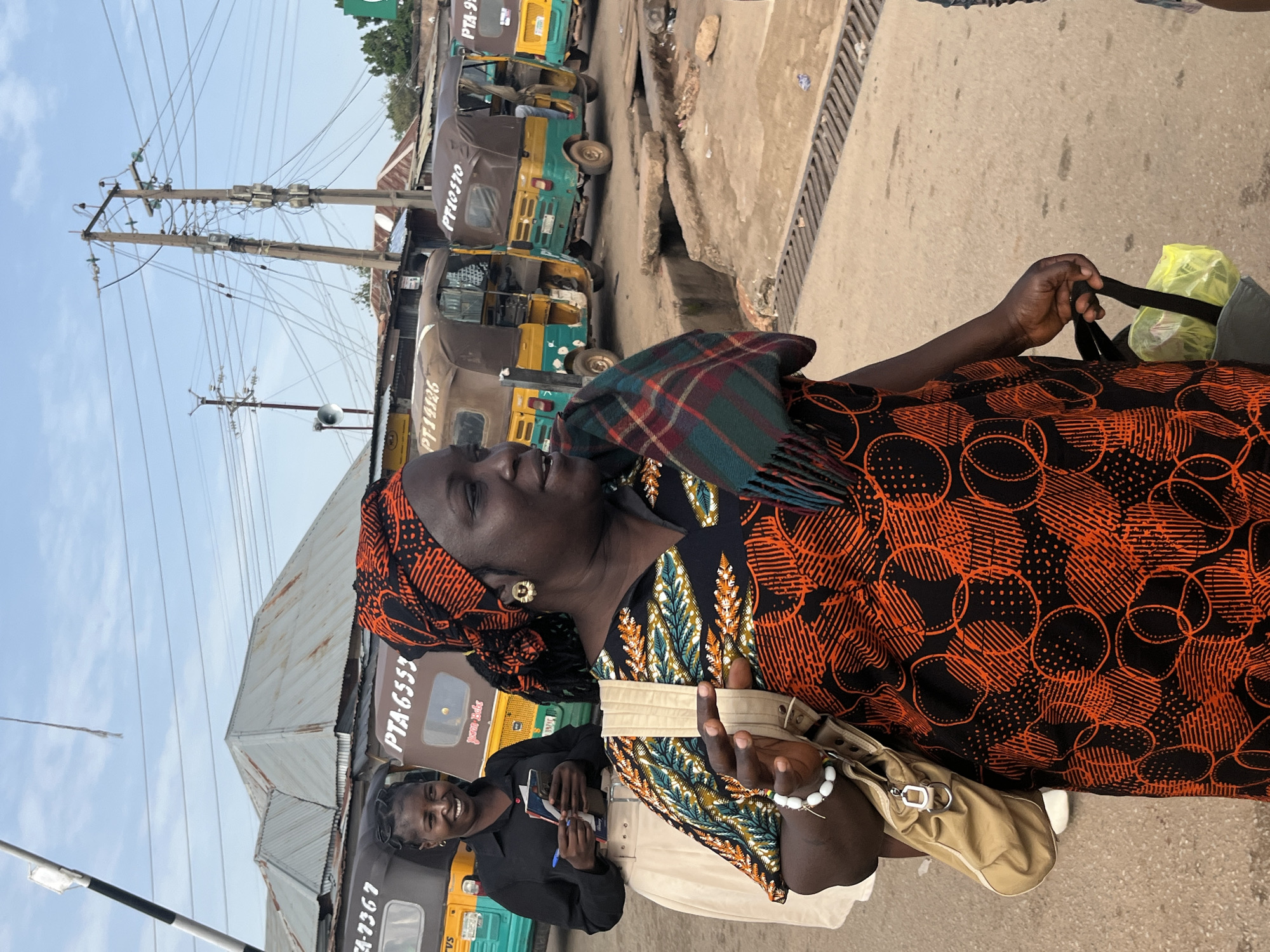 This media file captures a scene of daily life in Gura Top, Nigeria, featuring several people and the bustling environment of what appears to be a transportation hub or market. The primary subject is a woman in the foreground, adorned in a vibrant orange and black patterned dress and a headscarf. She is looking to her left, and her expression suggests contemplation or engagement in conversation. She holds a dark strap in her hand, possibly from a bag, and wears a bracelet.
Behind her, another woman is visible, smiling and holding some papers and a pen. This suggests an interaction, perhaps related to transportation services or a transaction. The background is dominated by several auto-rickshaws, commonly known as "Keke Napep" in Nigeria. These are parked in a line, indicating a waiting area for passengers or a commercial zone. Some of the auto-rickshaws have visible license plates or identifying numbers, with text such as "PTA-6553", "PT-14826", and "PF10570" clearly legible. Another partial text reads "yason Babe".
The setting appears to be outdoors, with a paved ground surface and a drainage grate on the right side. Overhead, there are prominent power lines and utility poles, indicating an urban or semi-urban environment. The lighting suggests it is daytime, likely morning or afternoon, with a bright, clear sky. The overall atmosphere conveyed is one of everyday activity, commerce, and local transportation.