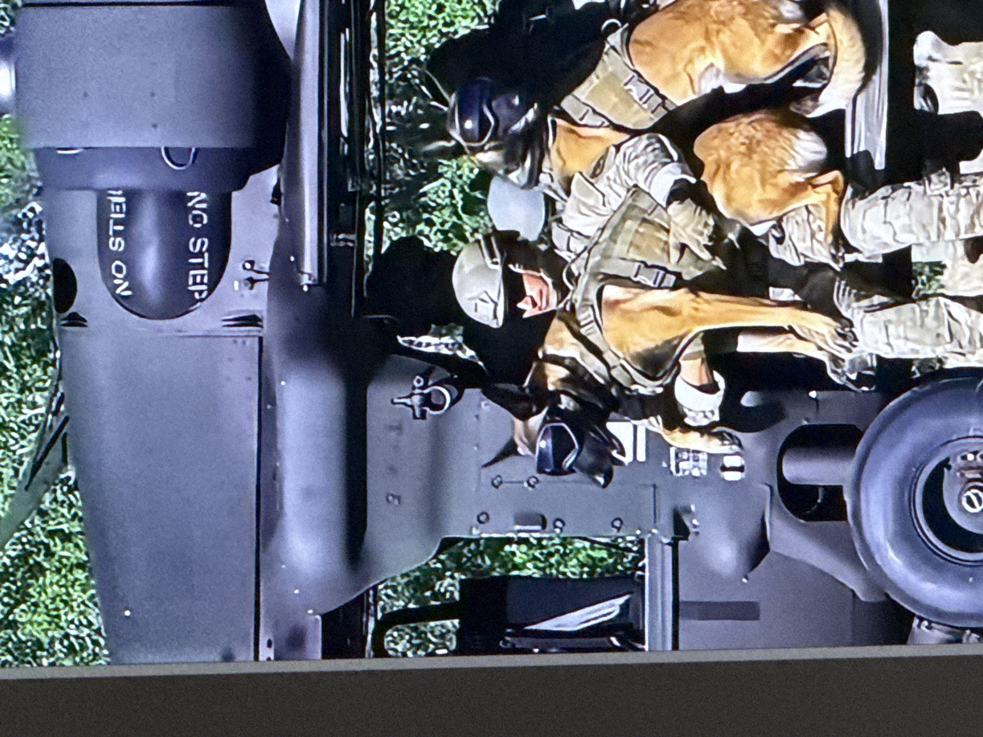 The image shows two military personnel and two dogs. The personnel are wearing camouflage uniforms, tactical vests, and helmets. One dog is a German Shepherd wearing goggles, and the other appears to be a Belgian Malinois. Both dogs are also wearing tactical vests. They are sitting on the back of a large, dark-colored vehicle, which appears to be a military transport. The vehicle has the words "NO STEP" and "NO STEIL" stenciled on it. The background is blurry, but appears to be green foliage, suggesting an outdoor setting. There are no visible landmarks that specifically place the scene in the city of Cahul, Moldova.