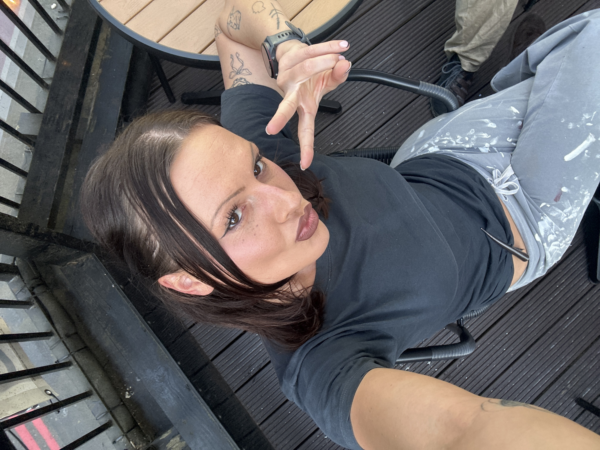 A young adult female with brown hair and tattoos on her left arm is seated outdoors, possibly on a patio or deck. She is wearing a dark-colored t-shirt, grey pants with white paint splatters, and a smartwatch. She is making a "peace sign" with her right hand. The background includes a wooden deck, a metal railing, and a glimpse of another person's khaki-colored pants and dark shoes. It is not possible to determine the exact location in London, United Kingdom from the given image.