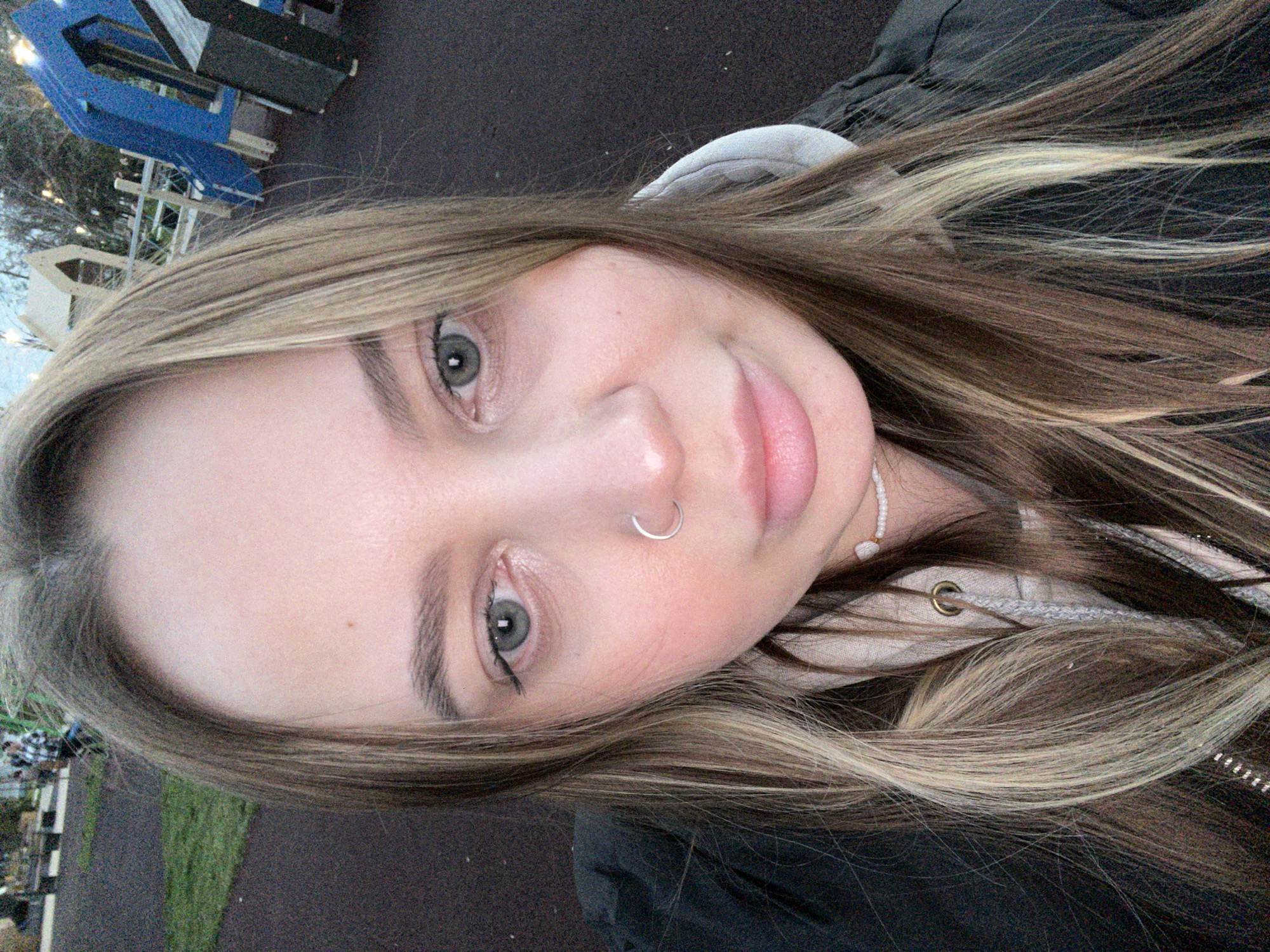 A selfie photograph shows a woman with light brown and blonde hair, light skin, and grey eyes. She has a silver nose ring in her left nostril. She is wearing a dark-colored jacket and a light-colored hooded sweatshirt. A white beaded necklace is visible. The background includes a playground with blue and gray structures, a paved surface, and a grassy area. The photograph may have been taken in Novopokrovskaya, Russia, but this cannot be confirmed from the image alone.