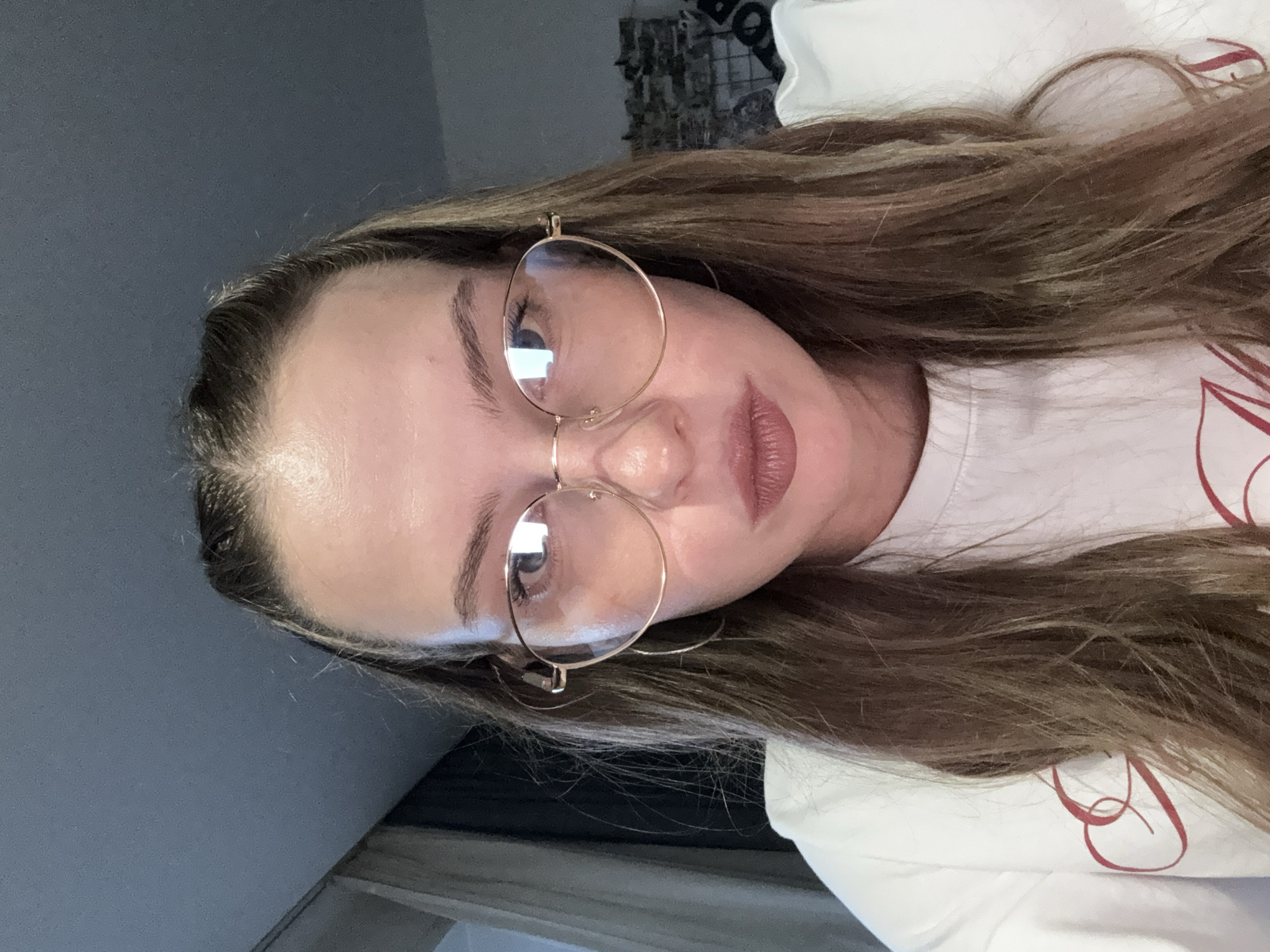 A young woman is shown from the shoulders up, with her head tilted to the left. She is wearing large, round, gold-rimmed glasses and has long, wavy, light brown hair. Her eyes are dark, and her lips are painted a soft pink. The background is out of focus, but appears to be an indoor setting with a plain wall to the left and what looks like a white t-shirt with a red cursive design on the right. The lighting is bright, suggesting it could be daytime, and there are reflections visible in her glasses. The overall impression is casual and relaxed.