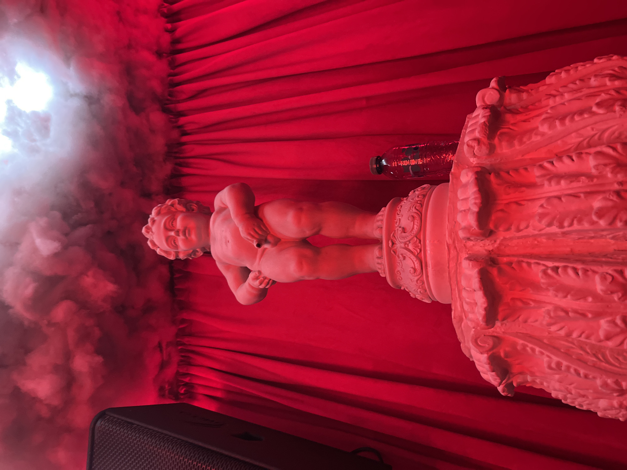 Here's a detailed description of the image:
**General Description:** The image shows a statue of a nude cherubic boy on an ornate pedestal, set against a backdrop of red fabric and a cloudy/smoky effect. The statue and its setting are illuminated with a red light.
**Objects:**
* Statue: A white or light-colored statue of a nude boy. The statue has a classical style, with intricate details.
* Pedestal: The statue stands on an ornate pedestal, also light-colored. The pedestal features carvings and decorative elements.
* Red Fabric: The background is draped with red fabric, which is folded to create vertical lines.
* Water bottle: a plastic water bottle is leaning on the statue's pedestal.
* Smoky Effect: The left side of the image features a hazy, smoky effect, possibly created by a fog machine.
* Speaker: the base of the image includes a black mesh speaker.
**Environment:**
* Lighting: The scene is lit with a strong red light, giving the entire image a monochromatic red tone.
* Setting: The presence of the red fabric suggests that the scene may be part of a stage, display, or a decorated interior space.
**Location:** The location given, San Bernardino Tlaxcalancingo, Mexico. is not directly evident in the image itself. However, it suggests that the scene is located at this place.
**Summary:** The image depicts a staged or decorative scene with a classical statue as the focal point, set against a red backdrop and a smoky effect. The red lighting adds a dramatic atmosphere to the image.