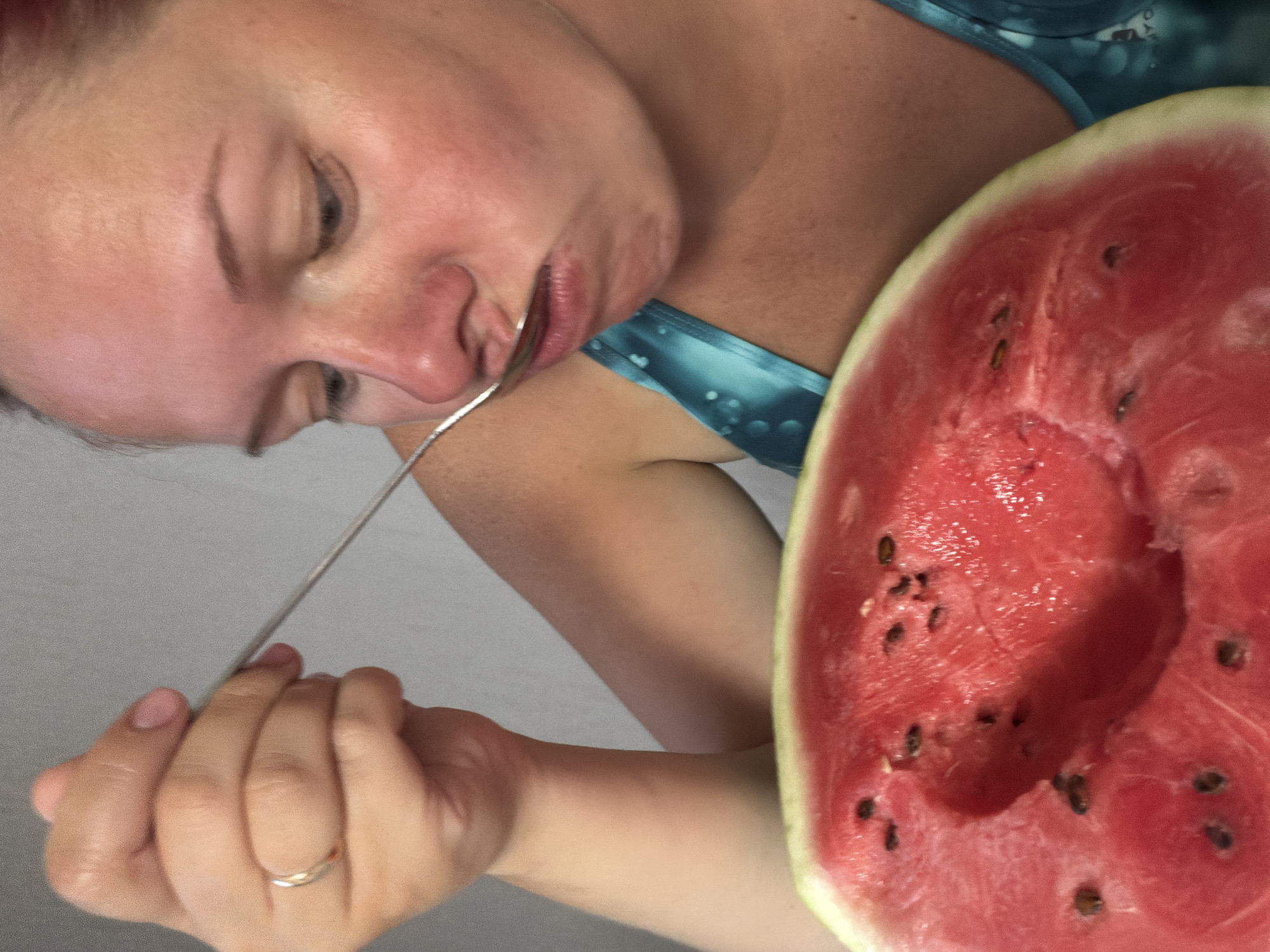 A woman is eating watermelon with a spoon. She is leaning over a cut half of a watermelon. The watermelon is pink and has black seeds. The woman is wearing a blue tank top. The background is a plain gray wall. The image was taken in Chaikovsky, Russia.