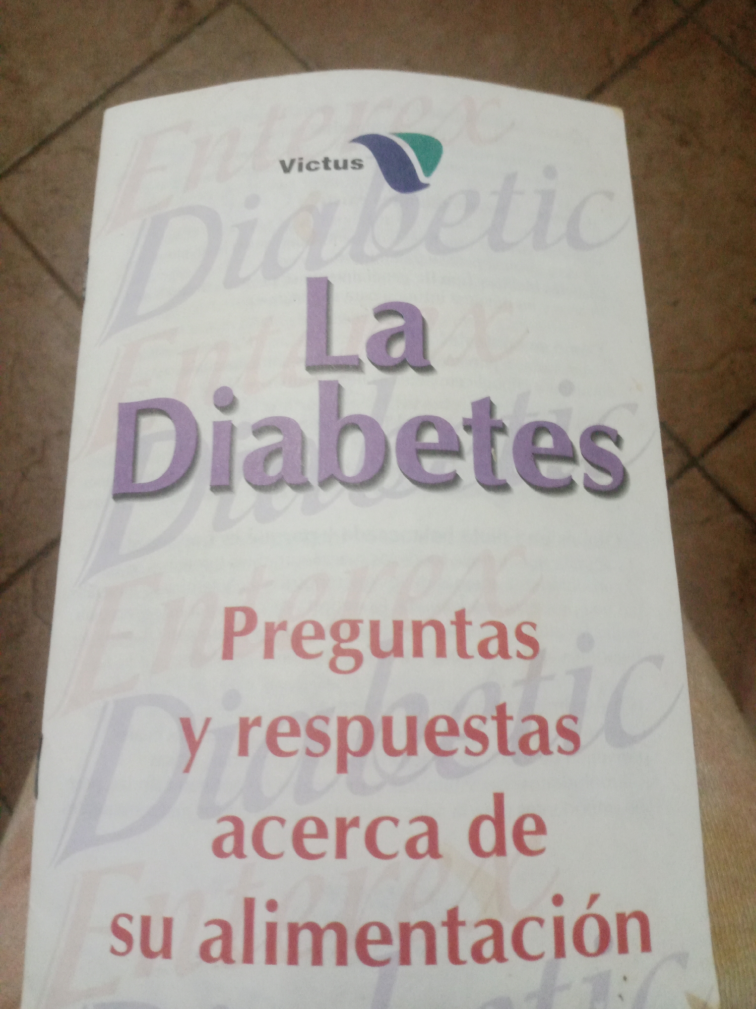 The image is a close-up shot of a book cover, titled "La Diabetes". The title is prominently displayed in large, purple, 3D lettering. Above the title, the word "victus" is visible, with a stylized blue and green logo next to it. Below the main title, the text "Preguntas y respuestas acerca de su alimentación" is printed in red. The background of the book cover features a faded, repeating pattern of text. The book is placed on a textured surface, possibly a floor or a fabric, with diagonal lines visible in the upper portion of the image. There is no indication of the location being Santa Ana, Venezuela.