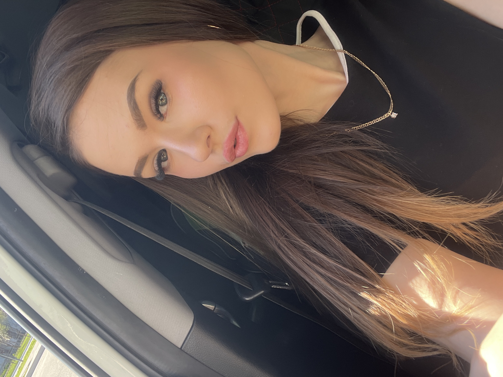 A young woman with long brown hair and light-colored eyes is reclining against the interior of what appears to be a car. She is wearing a black top with a white collar and a delicate gold necklace. Her makeup is visible, including eyelashes, eyebrows, and lip color. The car interior is visible, including the door panel and a seatbelt. The location of the scene cannot be determined from this image. There is nothing to suggest the location is the city of Chaikovsky, Russia.