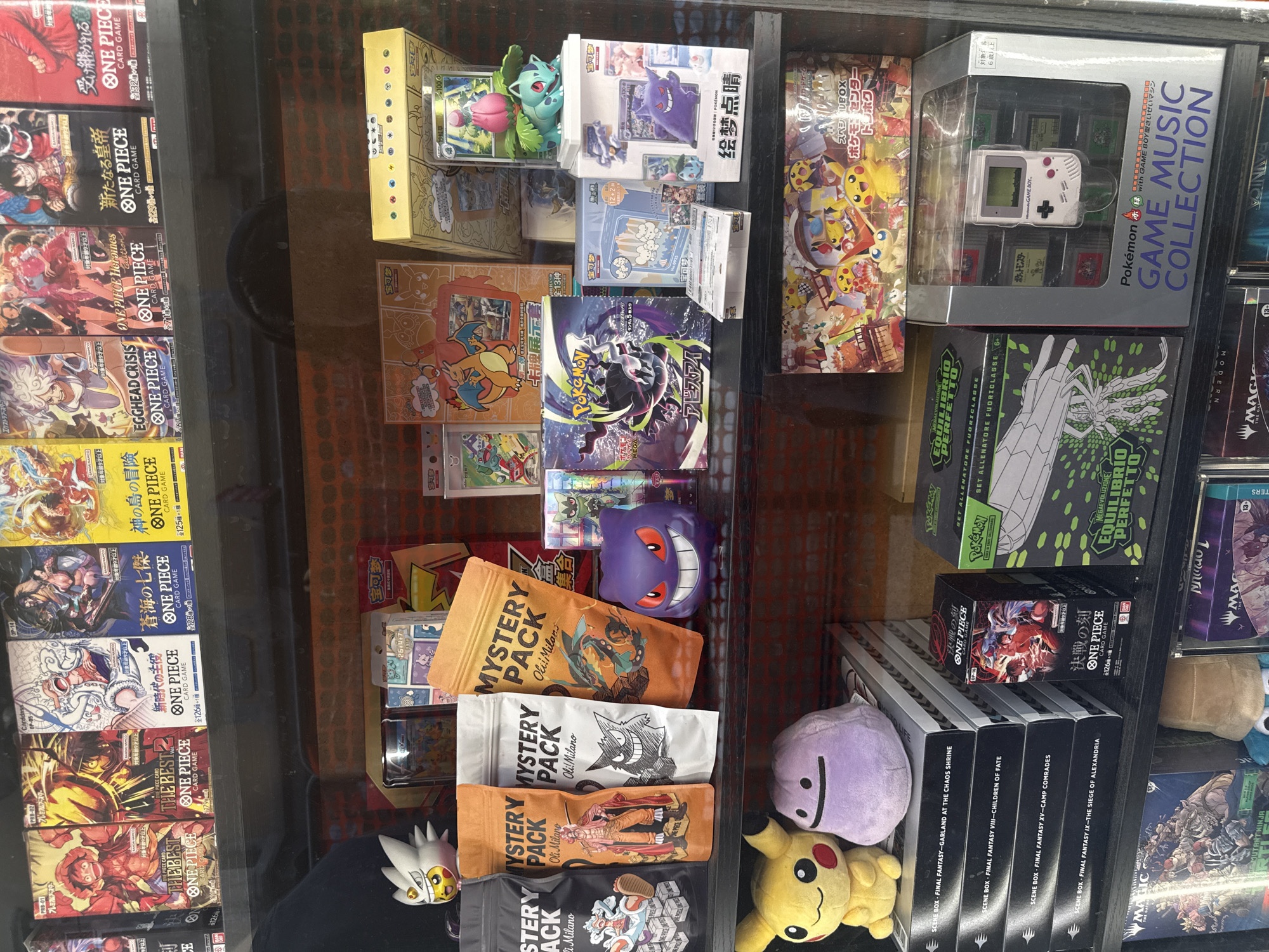 The image shows a shelf display of various collectible card games, toys, and merchandise. The items are predominantly from popular franchises like One Piece and Pokémon. There are stacks of card game booster boxes, collectible figures, and plush toys. Notably, there are several One Piece card game booster boxes with Japanese text and illustrations of characters. Several Pokémon-themed items are also visible, including figures, plush toys, and card game sets. One item is a "Pokémon GAME BOY MUSIC COLLECTION" which includes a replica Game Boy. The shelves are dark, and the display is behind glass, suggesting it's a retail environment. The presence of Japanese text on many of the products indicates that these are likely imported goods. The image does not provide any specific information about its location in Milan, Italy.