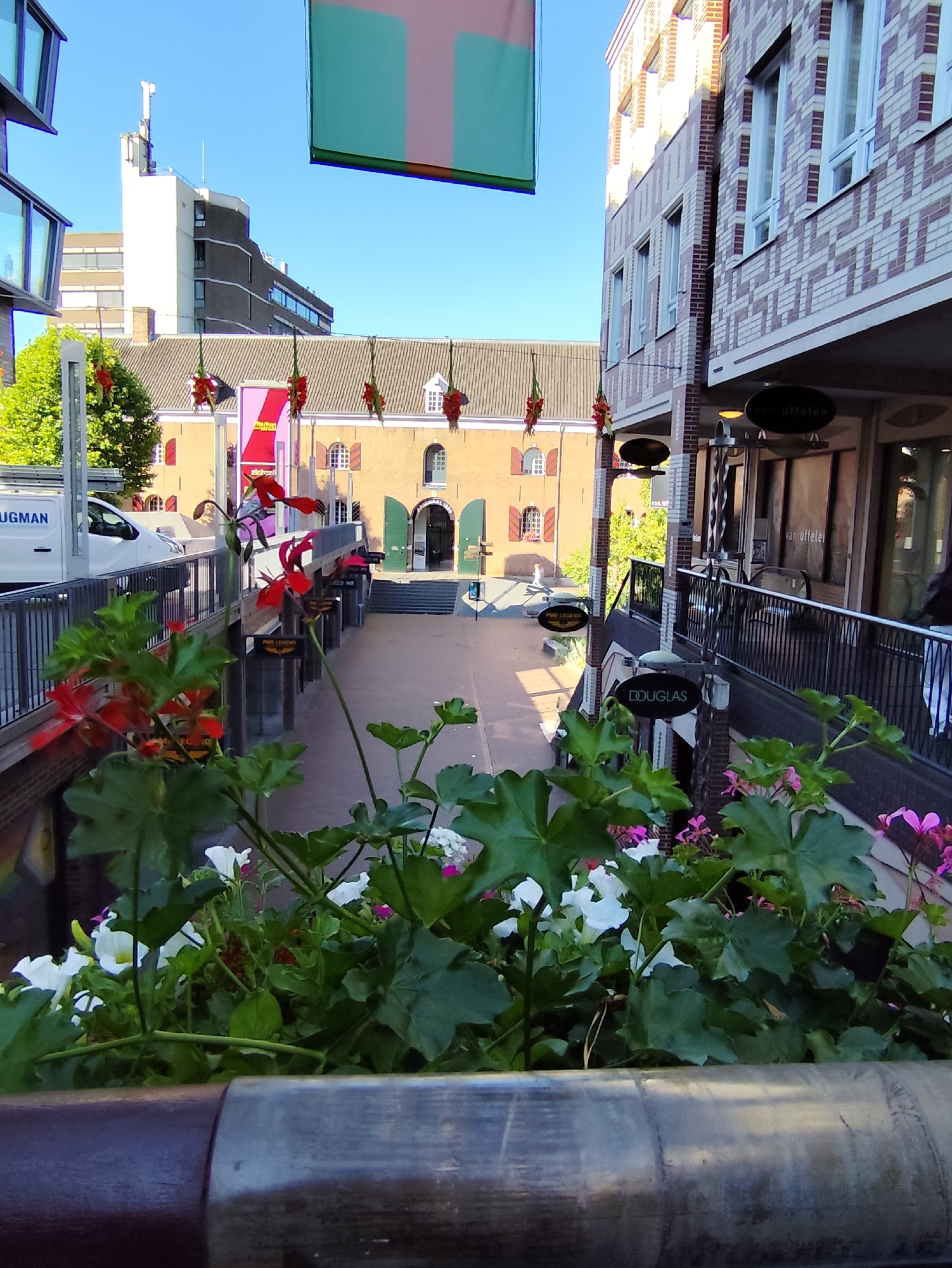 The image captures a street view in the city of Nijmegen, Netherlands, specifically near the Molenstraat. In the foreground, a planter box overflows with vibrant green foliage and a mix of red and white flowers, some with pink accents. Beyond the planter, a metal railing runs along a walkway, bordered by buildings on the right. These buildings feature a patterned brick facade in shades of beige and dark red, with tall, white-framed windows and awnings over storefronts. Signs for "van Uffelen" and "DOUGLAS" are visible.
The walkway leads to a courtyard area with more buildings. On the left, a white van with the partial text "UGMAN" is parked. Further back, a light brown building with a grey tiled roof and red-shuttered windows stands prominently. A green flag, split vertically into three sections of green and pink, hangs from a structure above. The sky is clear and blue. In the distance, modern buildings and a tall antenna are visible. The overall scene suggests a bright, sunny day in a European city, with a blend of historic and modern architecture.