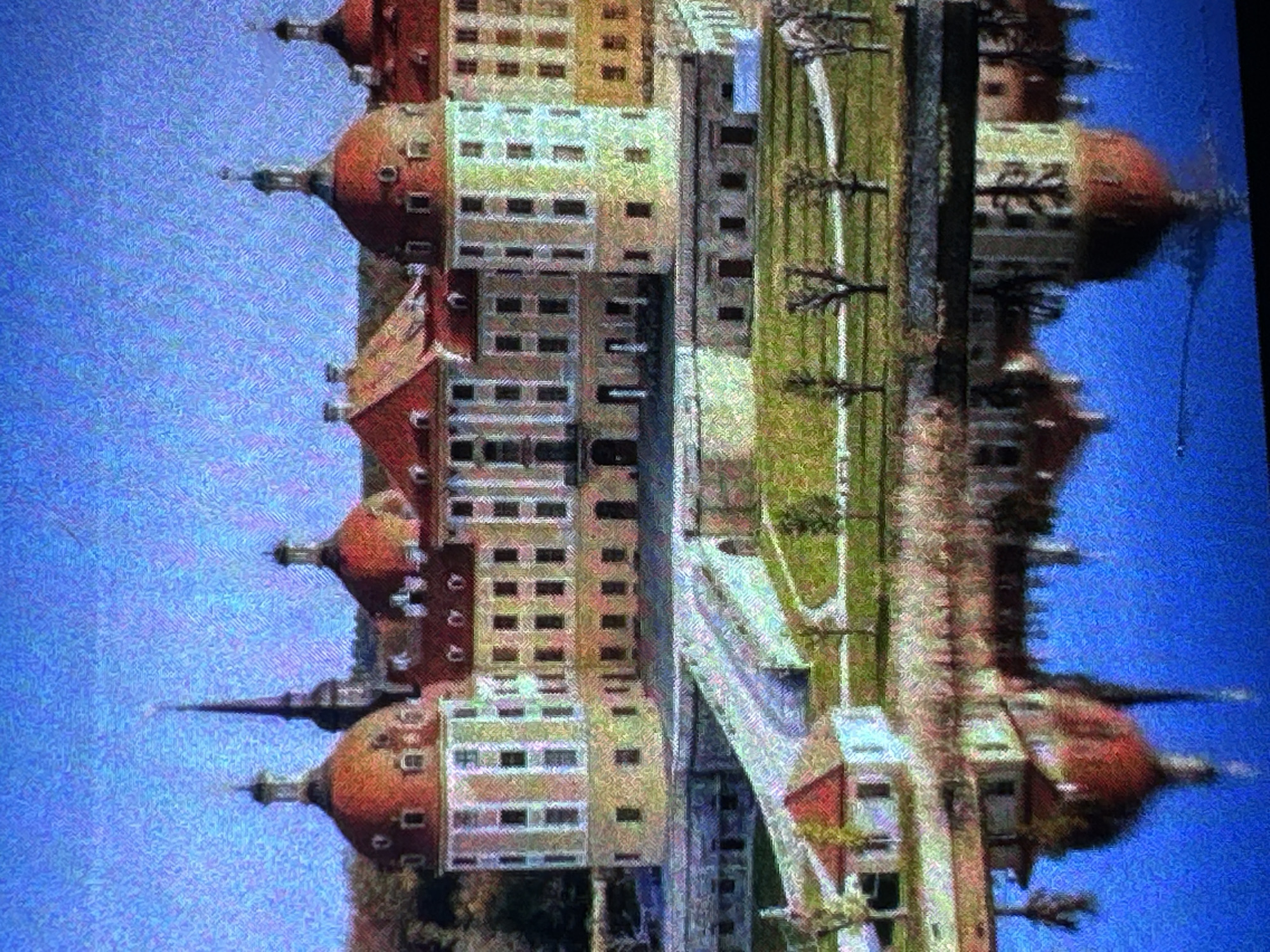 The image shows a picturesque view of a grand castle, likely a historical landmark, situated in a serene landscape. The castle features a striking architectural style with multiple red-domed towers and a central spire. It is surrounded by well-maintained grounds with green lawns, trees, and a body of water in the foreground. The water reflects the castle and the sky, creating a symmetrical and tranquil scene. The sky is a clear blue, suggesting pleasant weather. The overall impression is one of majesty and natural beauty. The image quality is affected by noticeable digital noise or grain, which gives it a slightly textured appearance.