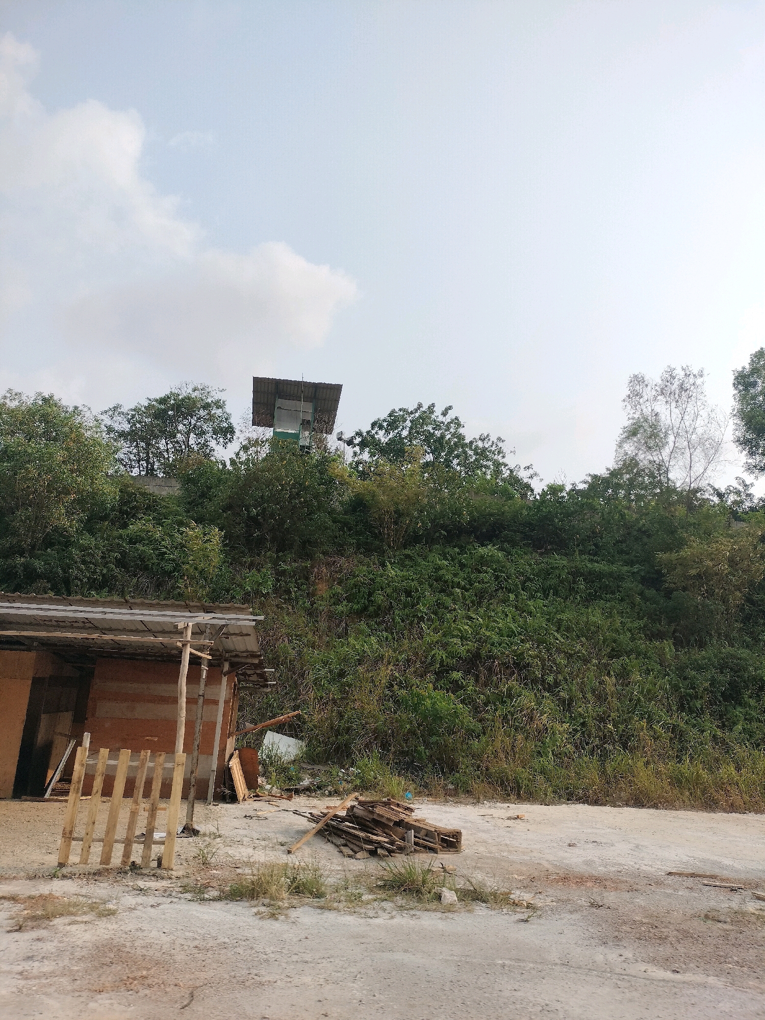 The image depicts an outdoor scene, likely in Batam, Indonesia, with a focus on a constructed area and a vegetated hillside. In the foreground, a rough, dirt and gravel surface is visible, with scattered piles of wooden planks and pallets. To the left, a simple, open-sided wooden structure with a corrugated metal roof is present, appearing to be under construction or in disuse. Behind this structure, a steep, densely vegetated hillside rises, covered in various types of green foliage and trees. On the crest of the hill, a small, elevated structure with a metal roof and walls, possibly a lookout post or utility shed, is visible, surrounded by trees. The sky above is overcast with diffused light. The overall impression is of an undeveloped or industrial area adjacent to a natural landscape. There are no people visible in the image.