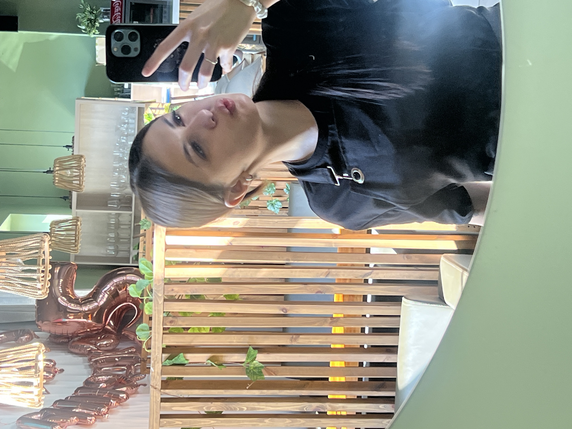 An indoor mirror selfie captures a young woman in a commercial establishment located in Красные Баки, Russia. The woman, positioned centrally, holds a black smartphone with a triple-lens camera in her right hand, extended towards the mirror. She has light-colored hair, with some strands pulled back and others falling over her left shoulder. She is wearing a black short-sleeved top or apron featuring a metallic D-ring and grommet detail on the left chest area. She wears a silver-colored ring on her left index finger, silver hoop earrings, and a white scrunchie on her left wrist. Her lips are pursed.
The background, visible in the reflection, features a light sage green wall. A wooden slatted partition with sparse green foliage (possibly ivy) runs across the mid-ground. Behind the partition, rose gold-colored foil balloons forming the numbers "2" and "5" are partially visible. Overhead, several pendant light fixtures with woven rope shades hang from the ceiling. To the right of the balloons, white wooden shelving units display multiple rows of clear drinking glasses. Further right, a red and white "Coca-Cola" sign is discernible, along with a dark wall-mounted speaker. In the lower-left background, a segment of a light-colored, upholstered booth or seating is visible.