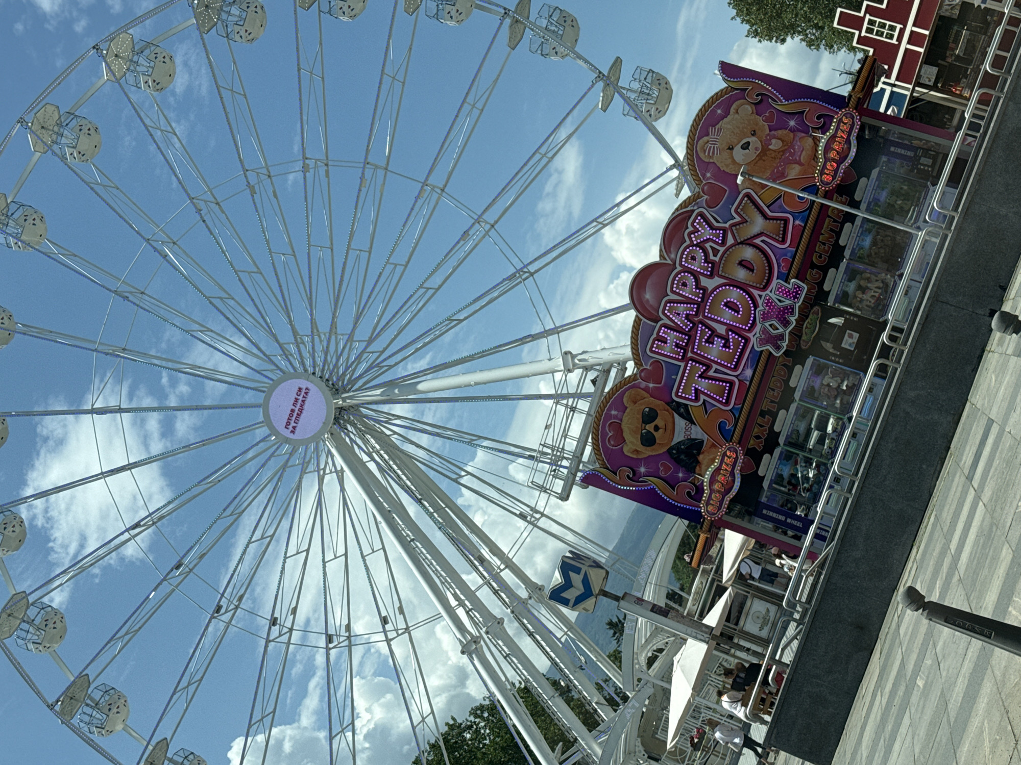 This image depicts a sunny outdoor scene in Sofia, Bulgaria, centered around a large white Ferris wheel. The Ferris wheel dominates the upper portion of the frame, set against a bright blue sky with scattered white cumulus clouds. Its central hub displays an illuminated circular sign with the Bulgarian Cyrillic text "ГОТОВ ЛИ СИ ЗА ГЛЕДКАТА?", translating to "ARE YOU READY FOR THE VIEW?". The spokes of the Ferris wheel feature small blue integrated lights, and its passenger gondolas are white with perforated details.
In the mid-ground, a brightly decorated amusement game stand, "HAPPY TEDDY XXL TEDDY WINNING CENTRE," is prominently displayed. The stand is adorned with neon-style lettering and large cartoon graphics of teddy bears, one in sunglasses and a "BOSS" jacket, and another in a pink dress. It advertises "BIG PRIZES" and features multiple glass display cases showcasing plush toys. To the left of the game stand, several white outdoor umbrellas shade a seating area with tables, where a few casually dressed individuals are observed walking or sitting. A blue and white sign with an "M" logo, indicative of the Sofia Metro, is partially visible beneath the Ferris wheel's structure.
The foreground consists of a paved public space with a pattern of light and dark grey rectangular tiles. A dark bollard is visible in the lower-left. In the background, beyond the urban elements, green trees frame the scene, and a distant mountain range, characteristic of Vitosha Mountain, is visible under the clear sky. The scene portrays a recreational area within the city.