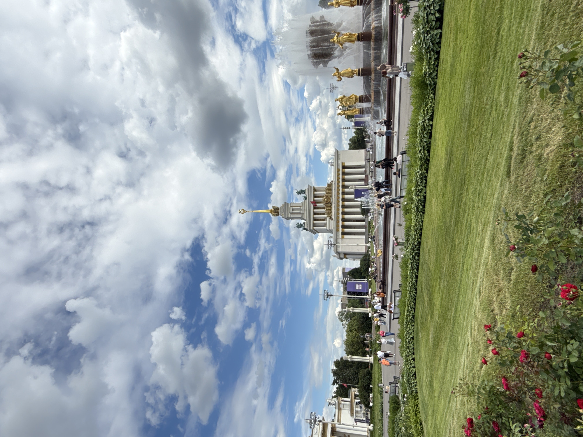 The image captures a panoramic view of the Friendship of Peoples fountain at VDNKh (Exhibition of Achievements of National Economy) in Moscow, Russia, on a day with partly cloudy skies. The iconic fountain, adorned with gilded sculptures of women representing Soviet republics, is the central focus, with water spraying high into the air. Behind the fountain, a grand building with neoclassical architecture, featuring a tall spire topped with a golden star, dominates the background. Several people are visible, some walking and others sitting on benches, enjoying the expansive green lawn and manicured bushes in the foreground. The scene is set in a public outdoor space, likely a park or exhibition grounds, with trees and additional structures suggesting a well-developed urban environment.