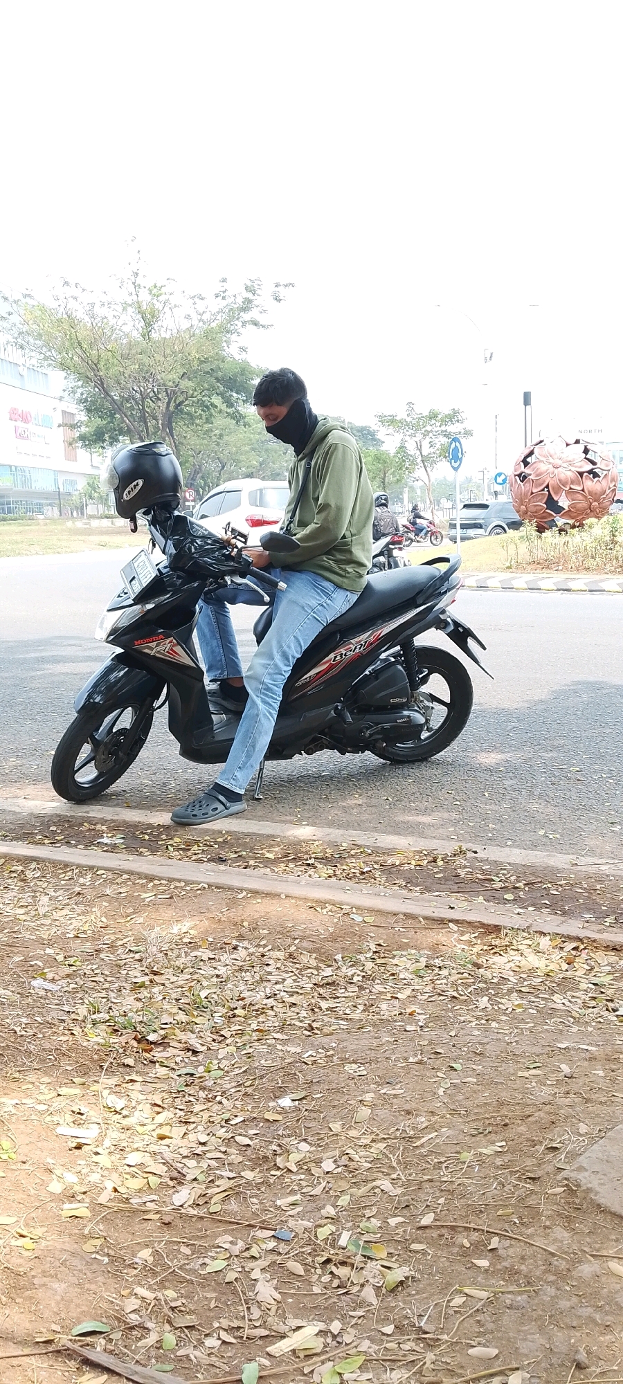 A person is sitting on a black motorcycle with a black helmet resting on its handlebars. The person is wearing a light green hooded jacket, blue jeans, a black face mask, and dark gray clogs. The motorcycle is parked on the side of a road in Gunung Putri, Indonesia, with trees and buildings visible in the background. A large, spherical art installation with a floral pattern is present to the right of the motorcycle. The ground in the foreground is covered with dry leaves and twigs.