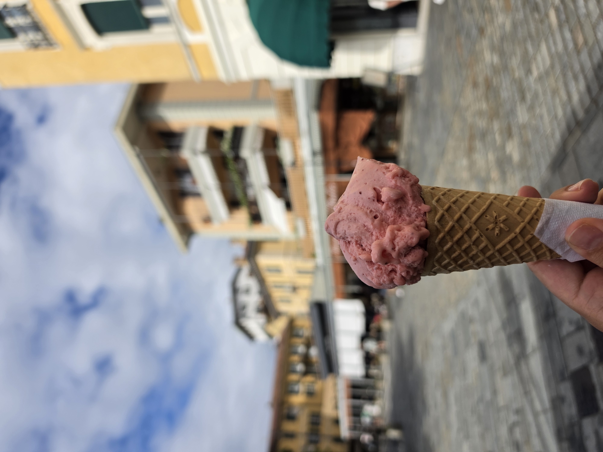 In the foreground, a hand holds a strawberry gelato cone with a waffle cone and white paper wrapper. The background shows a street in San Giovanni, Italy, with buildings and a cloudy sky. The buildings are colorful, with yellow and orange facades and balconies. There are shops with awnings and people walking in the distance. The ground is paved with cobblestones.
