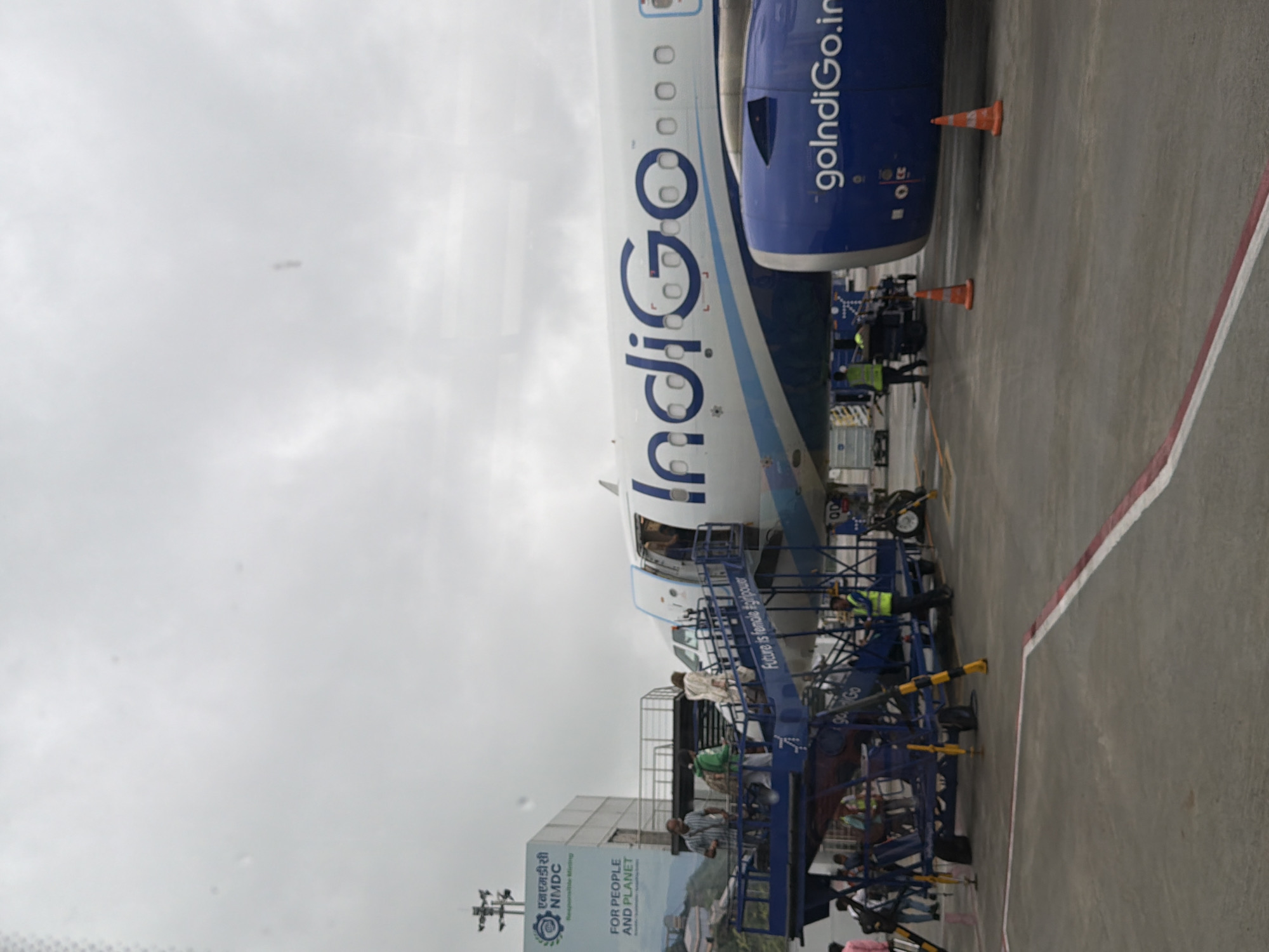 The image depicts an Indigo Airlines airplane parked at an airport, likely Rajiv Gandhi International Airport in Hyderabad, India, based on the presence of an NMDC advertisement which is a public sector undertaking headquartered in Hyderabad. Passengers are seen disembarking or boarding the aircraft via a mobile staircase. The sky is overcast. In the foreground, there is an airport tarmac with markings and a baggage cart. An Indigo Airlines logo is visible on the aircraft's fuselage, along with the website address "goindigo.in". There are also several orange traffic cones placed on the tarmac. The overall scene is that of an active airport environment during daytime.