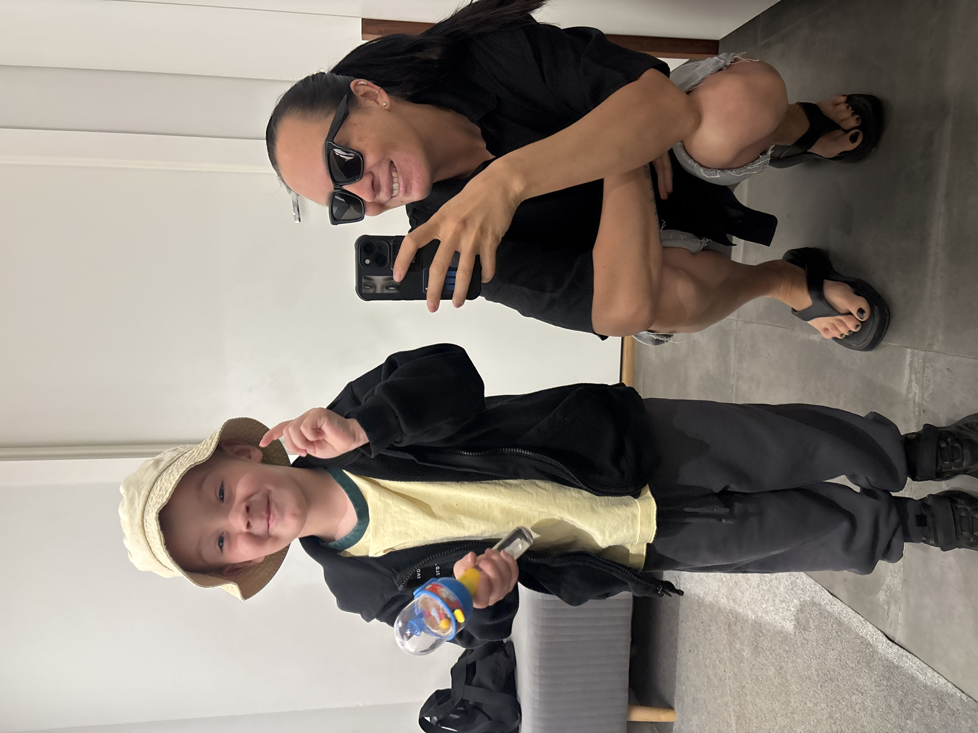 A selfie taken in Chaikovsky, Russia, shows a woman and a child, likely in a mirror. The woman squats, wearing sunglasses, a black top, and denim shorts, holding a smartphone. The child stands in front of the woman, wearing a bucket hat, a yellow t-shirt under a black zip-up jacket, and grey pants, holding a toy. The background is a simple white wall and grey flooring.