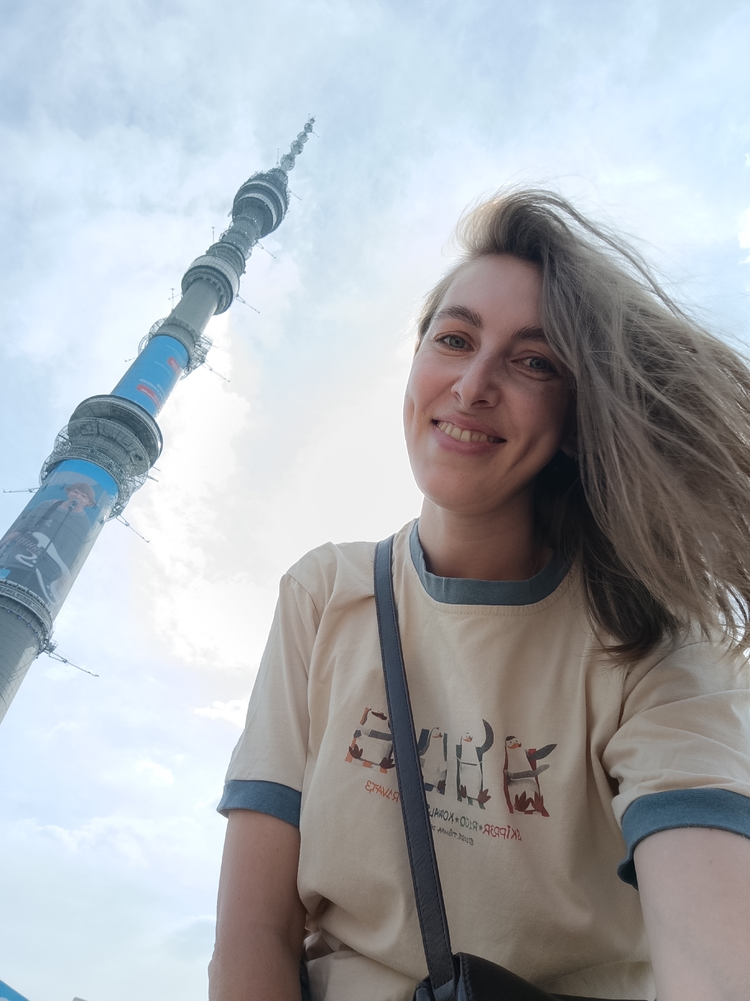 The image captures an adult female smiling in a low-angle selfie, with the Ostankino Tower in Moscow, Russia, prominently featured in the background. The woman, positioned in the lower right portion of the frame, has light brown, windblown hair and light-colored eyes, displaying dimples as she smiles directly at the camera. She wears a light beige ringer t-shirt with blue-grey trim on the collar and sleeve cuffs. The t-shirt's front is adorned with a graphic depicting six stylized penguins. A dark strap, likely from a shoulder bag, is visible across her right side. Behind her, the massive grey cylindrical structure of the Ostankino Tower ascends diagonally from the lower left towards the upper center, showcasing multiple platforms and tapering as it rises. Several large blue banners, featuring white text (including Cyrillic script) and an image of a person holding a guitar, are wrapped around various sections of the tower. The sky above is a bright, partly cloudy blue.