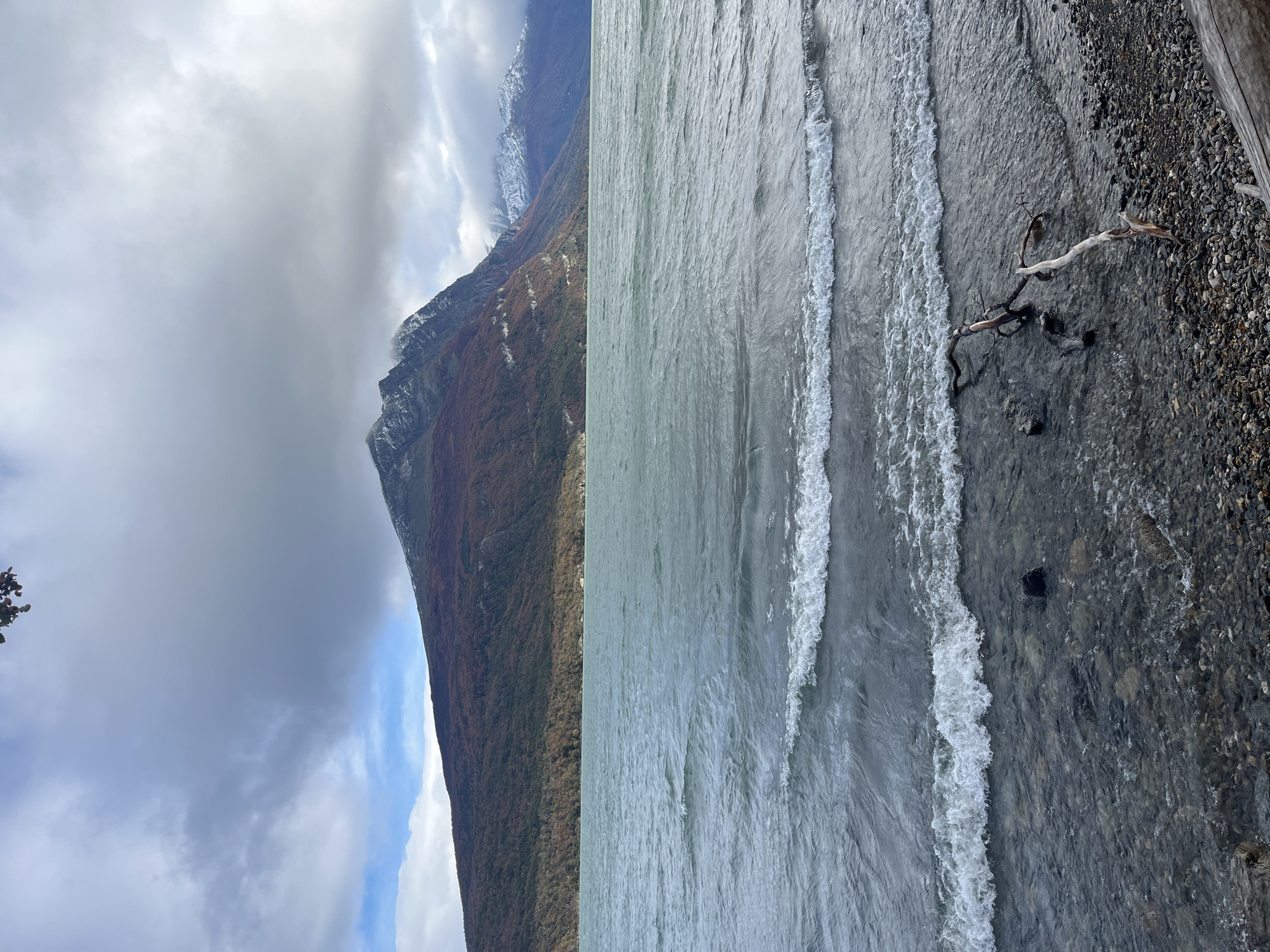 The image depicts a landscape with a mountain on the left side and water on the right side. The mountain is partly covered by clouds and has a mix of trees and rocky terrain. The water appears to be a lake or sea, with small waves reaching a rocky shore. There's driftwood on the shore. The location is suggested to be near Lapataia, Argentina.