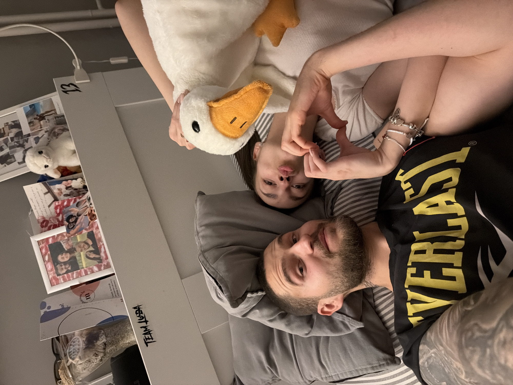 A man and a woman are lying on a bed with gray pillows and striped sheets. The man is on the bottom, wearing a black T-shirt with yellow lettering and has a short beard. The woman is above him, holding a white plush goose with an orange beak. Her hands are posed to make a heart shape. The background includes a gray wall, and on a shelf, there are photographs, a white stuffed cat, and the number "13" written on it. Additionally, the phrase "TEAM AGEBA" is visible. There are no indicators about the city of Sofia, Bulgaria.