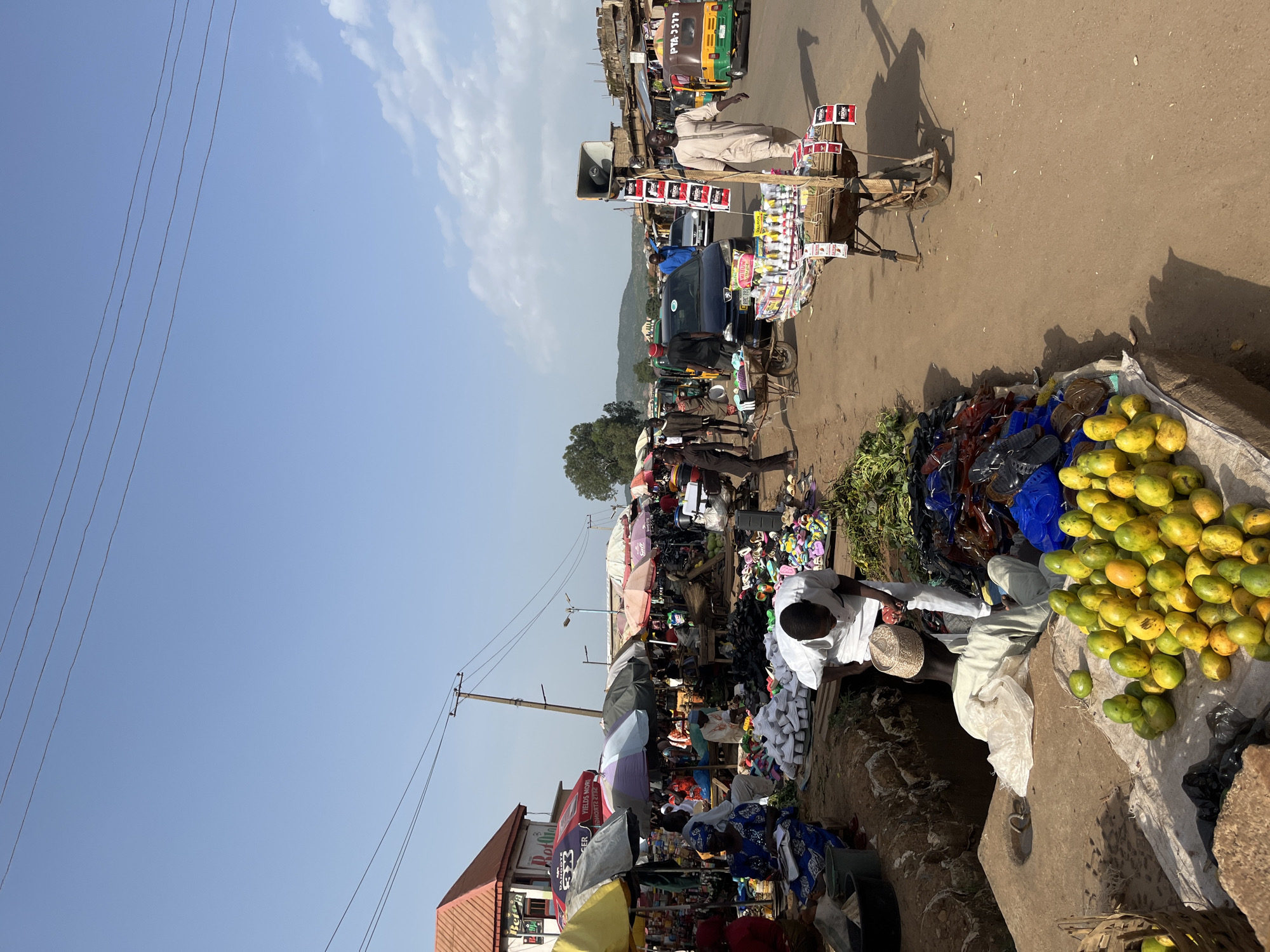 This media file captures a vibrant market scene in Rafin Par, Nigeria. The image is oriented vertically, showing a busy street lined with vendors and shoppers. The sun is high in the sky, suggesting it is daytime. The weather appears clear with a few wispy clouds visible.
The primary subjects are the people engaged in market activities. There are numerous individuals, some dressed in traditional attire, tending to their stalls, interacting with customers, or moving through the market. Several stalls display a variety of goods, including fruits (likely mangoes, as a large pile is prominently featured), footwear, and other miscellaneous items. A cart with a speaker is also visible, suggesting announcements or music might be part of the market atmosphere. Various vehicles, including a brightly colored auto-rickshaw, are present, indicating a transportation hub within or near the market.
Notable details include the dense arrangement of stalls and merchandise, creating a sense of lively commerce. The presence of electricity poles and wires suggests a connection to infrastructure, despite the rustic appearance of some stalls. Shadows cast by the sun indicate the time of day.
The overall emotion conveyed is one of bustling activity and community engagement. The setting is an outdoor market, likely a central gathering point for the local population.
Visible text includes "PTA-3577" on the auto-rickshaw and various logos and text on banners and products, although much of it is not clearly legible. One banner near the foreground appears to say "YIELD MORE ENERGY" and another has the word "SPRINGER".