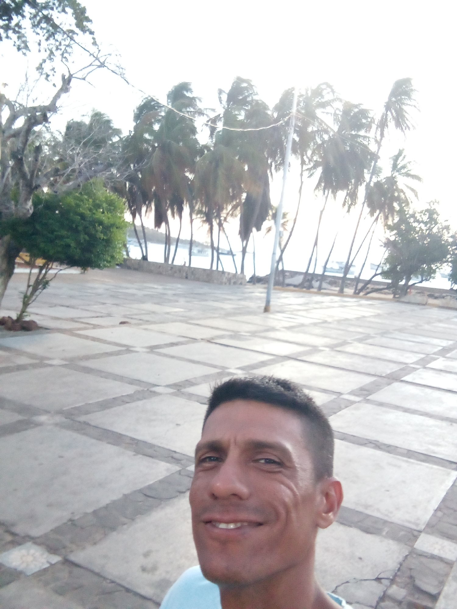 The image is a selfie taken by a man with short, dark hair, and a light blue shirt, smiling at the camera. He is likely standing in the Plaza of Juan Griego, Venezuela. The plaza is paved with a tiled pattern of squares. In the background, a line of palm trees are visible along the coast. The sun is either rising or setting, creating a bright, hazy effect.