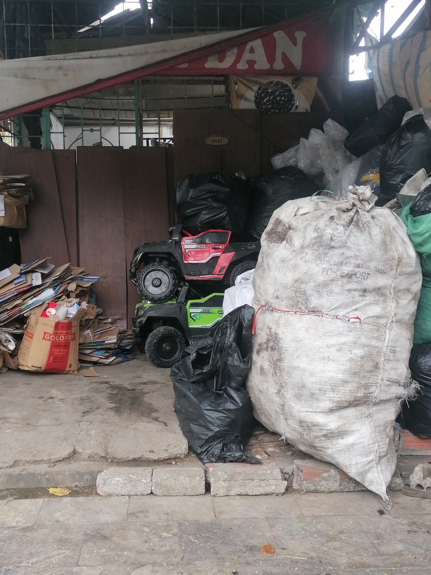 This media file captures a cluttered outdoor scene, likely a storage or waste collection area, situated in Bai Chay Ward, Vietnam. The overall impression is one of disarray and discarded items.
The central focus is a large, dirty white woven sack, appearing full and tied at the top. Partially visible black text on this sack includes "2680511, DAR ES SALAAM, TANZAN" and "FOR EXPORT", indicating its contents are destined for international shipment. Surrounding this sack, and extending to the right, are numerous black plastic garbage bags, piled high and bulging with refuse.
To the left of the large sack, two children's electric ride-on cars are stacked: a red one on top and a green one below. Both cars have "CHE" printed on their sides; the red car also shows "66", and the green one "G6". Further left, a substantial pile of flattened cardboard boxes and miscellaneous scrap paper is accumulated. At the bottom of this pile, a brown cardboard box from "GOLD FRY" is visible, featuring Vietnamese text that translates to "06 thực vật", "GOLD FRY", and "Chiên Vàng. Giòn Tan" (Golden Fry. Crispy), along with the number "#5271154".
The background consists of wooden panels or a brown wall, with a small white sign bearing the number "4042" attached to one panel. Above, a red banner with the white letters "DAN" is partially visible, and a metal grate structure suggests an enclosure or building facade. The foreground shows a worn concrete ground with cracks and some loose grey paving blocks. The lighting is soft, suggesting daytime, possibly overcast, and there are no people present in the image.