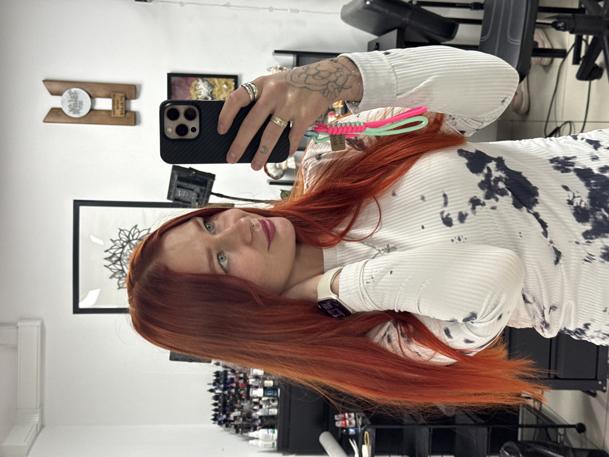 A person with red hair takes a selfie. The individual wears a white shirt with a black tie-dye pattern, an Apple Watch, and multiple rings. The background includes tattoo paraphernalia, framed art, and a salon chair. The location is possibly a tattoo studio but not determined in Rostov-on-Don, Russia.