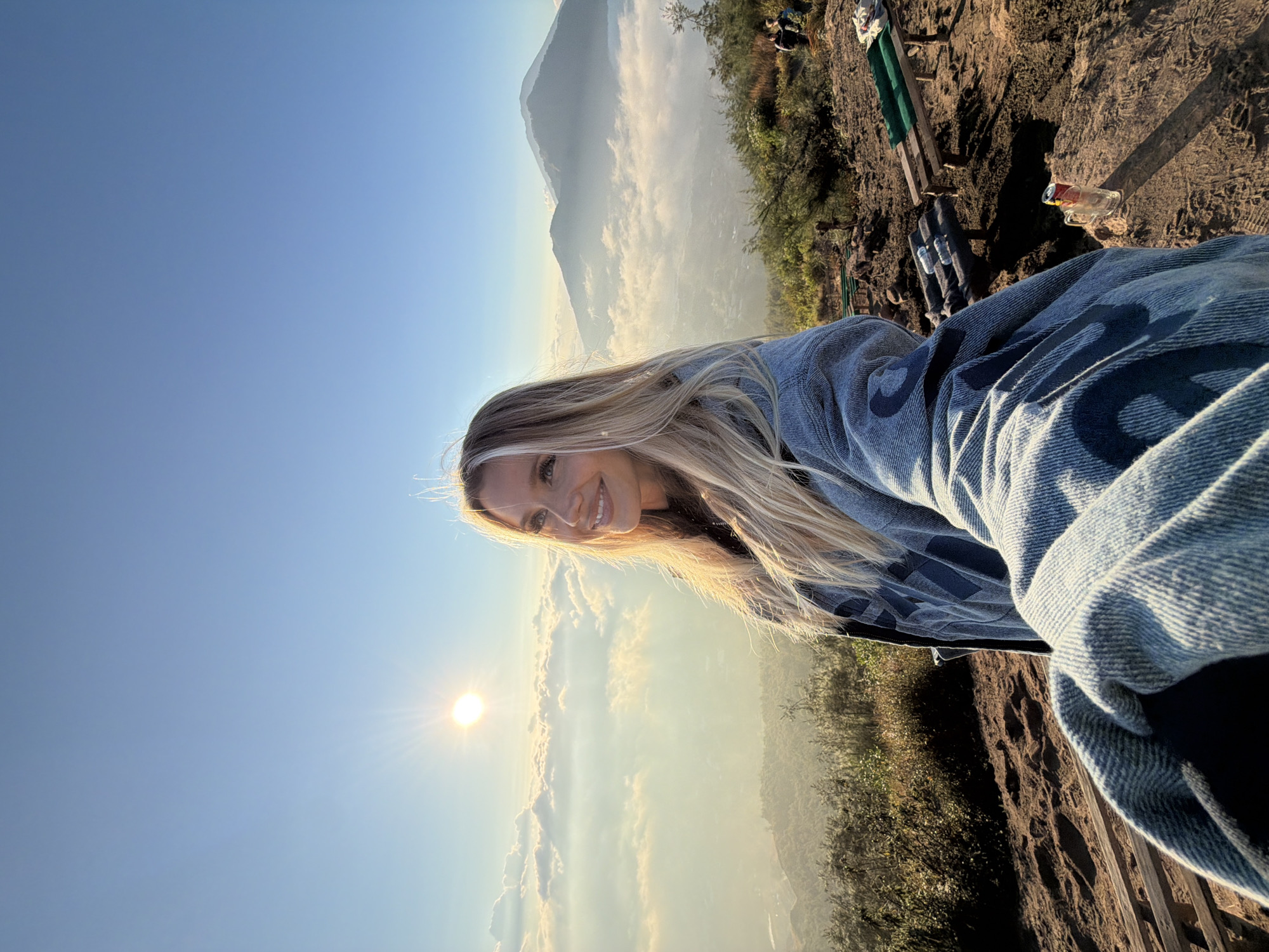 A woman with blonde hair smiles at the camera in a selfie taken from a high vantage point, likely a mountain summit, in Kintamani, Indonesia. The sun is rising, casting a warm glow and creating sun flares in the clear blue sky. Below, a sea of clouds stretches across the landscape, with the peaks of distant mountains emerging from the mist. The woman is wearing a denim jacket. In the foreground, the rocky, uneven terrain is visible, with some sparse vegetation. There are several wooden benches and a small table scattered around the area, suggesting it's a viewpoint for tourists. Two figures can be seen in the background on the right, also observing the sunrise.