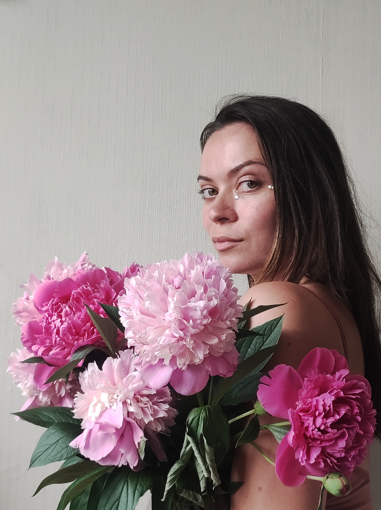 The image shows a woman holding a bouquet of pink peonies against a textured off-white background. The woman has long, dark hair and is looking over her shoulder towards the camera. She has makeup on and appears to be wearing a light-colored top. The peonies are large and full, with a mix of light and dark pink petals. The lighting suggests an indoor setting. The provided metadata does not indicate any specific location in Chaikovsky, Russia.