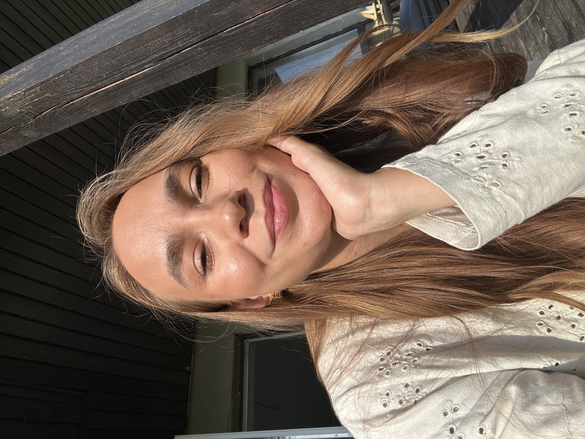 A young woman with long, auburn hair is captured in a close-up, selfie-style shot, oriented vertically. She is outdoors, with a wooden beam and dark wooden siding visible in the background, suggesting a porch or deck. Sunlight illuminates her face, casting a warm glow and highlighting her features. She has fair skin, light brown eyes, and her lips are slightly parted, wearing a subtle pink lipstick. Her left hand is resting gently on her cheek, and she is wearing a white, eyelet-embroidered top. A gold earring with a geometric design is visible in her left ear. The natural light and the relaxed pose suggest a pleasant, possibly sunny day. The image has a candid and intimate feel, with the woman appearing calm and content. No visible text is present in the image. The location context is Kharnavy, Russia.