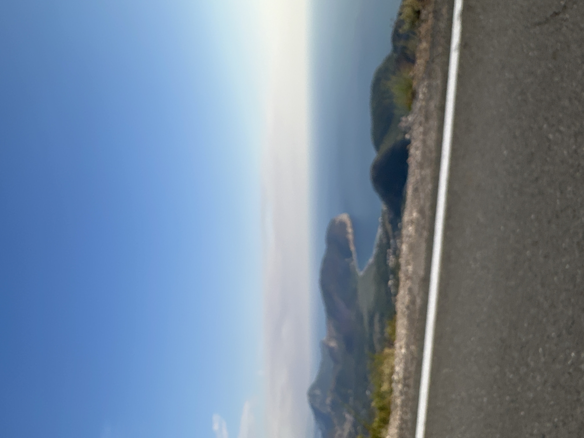 The image shows a slightly out-of-focus, elevated view of a coastal landscape. In the foreground, a gray asphalt road with a white line marking the edge is visible. The middle ground features rolling, green-covered hills that slope down towards the sea. A bay with calm, blue water is prominent, with a curved coastline. Further out, the sea meets a hazy horizon where the sky transitions from a bright, clear blue to a lighter, washed-out tone. The scene is devoid of people or any specific man-made structures apart from the road. The location is identified as being in the city of Novoselje, Montenegro, suggesting the view is from a vantage point overlooking the Adriatic Sea.