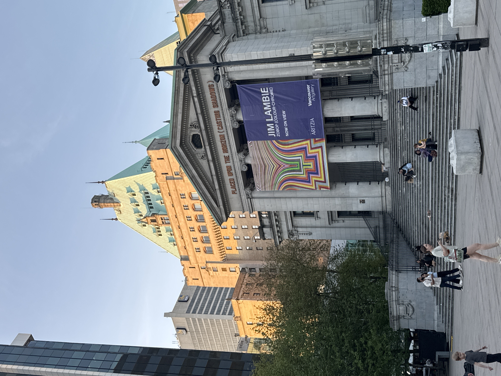 Here's a description of the image:
The image shows the Vancouver Art Gallery in Vancouver, Canada. A banner for a Jim Lambie exhibition (ZOBOP) hangs on the front of the building. People are milling around on the steps. Various buildings are visible behind the gallery. The sky is light blue.