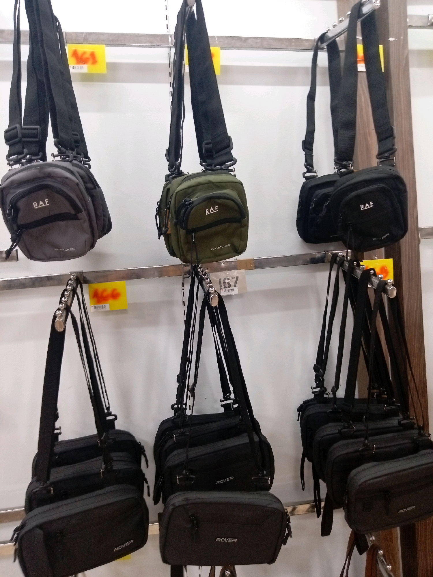 The image shows a retail display of shoulder bags in Batam, Indonesia. The bags are arranged on two metal shelves. On the top shelf, there are three bags: a gray bag on the left, a green bag in the middle, and a black bag on the right. All three bags on the top shelf have the brand "RAF" printed on them. On the bottom shelf, there are multiple black shoulder bags, with some displaying the brand "ROVER." Yellow price tags are visible on the shelves. The background is a plain white wall. The scene depicts a commercial setting, likely a store or a market stall, where these bags are being sold.