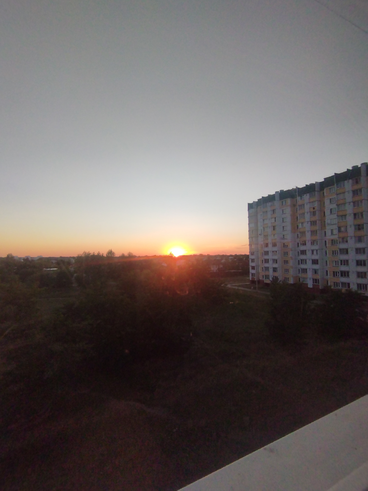 The image captures a view of a high-rise apartment building in Homyel, Belarus, during a sunset. The sky is a gradient of light grey at the top, transitioning to a soft orange and yellow near the horizon. The sun is visible as a bright, luminous orb on the left side of the image, casting a warm glow and creating lens flares.
The apartment building, a multi-story structure with a light-colored facade and dark roofline, dominates the right side of the frame. It appears to be an older, typical residential building found in Belarusian cities, with visible rows of windows. In the foreground and mid-ground, there is a dense expanse of dark foliage, likely trees and bushes, obscuring the ground level. In the distance, beyond the foliage and to the left of the apartment building, a low-rise cityscape is visible under the twilight sky. The overall scene suggests an urban or suburban environment observed from a vantage point, likely a window or balcony, looking towards the west. There are no people or specific actions depicted in the image.