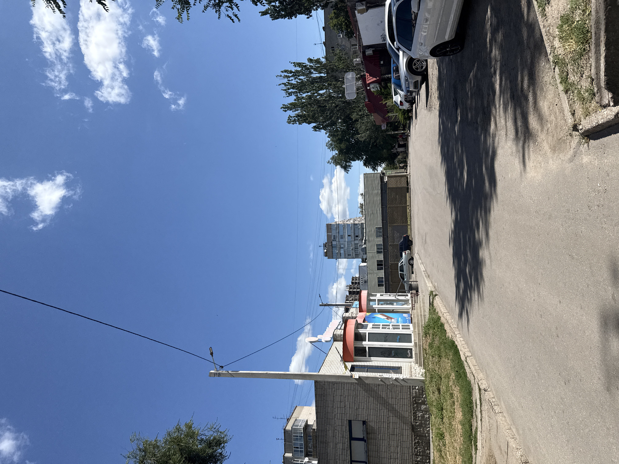 This is a street-level, eye-level, medium shot on a sunny day in Zaporizhzhia, Ukraine. The street is paved and appears to be in a commercial or residential area. On the left side of the street is a low-rise building with a brick facade and several windows. In front of this building are two glass-fronted storefronts with orange awnings above them. A tall, slender pole with a street light is positioned in front of these storefronts. The street is lined with parked cars on the right side, including several white sedans and SUVs. Beyond the parked cars, there are trees and other buildings, including a taller, multi-story building in the background. The sky is clear blue with scattered white clouds. The scene is quiet and still, with no visible people or activity.
