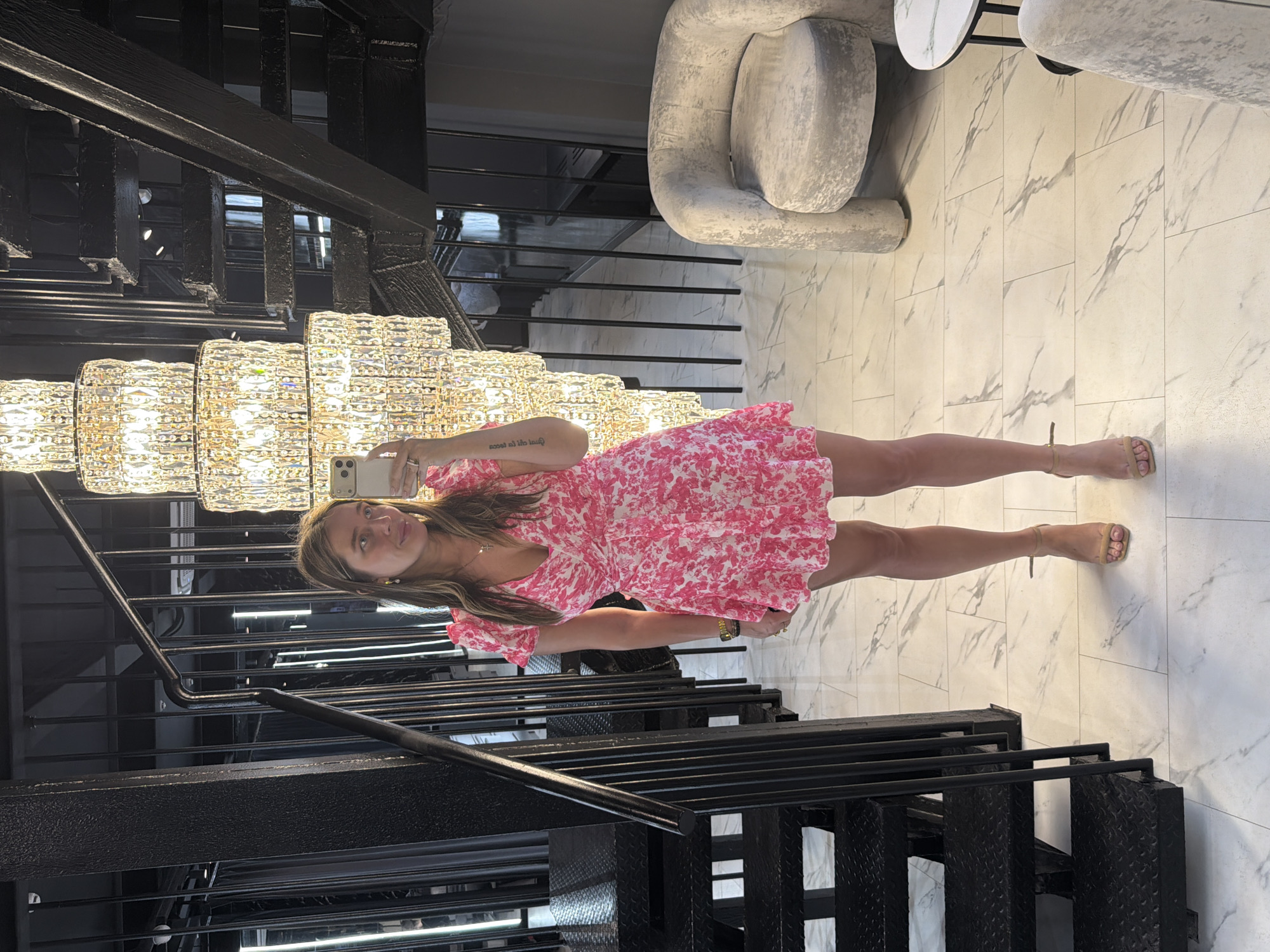 A young woman stands indoors, illuminated by a large chandelier, taking a selfie with her smartphone. She is wearing a short, pink floral dress and light brown heeled sandals. She has medium-length blonde hair and appears to be wearing a necklace. The setting appears to be a high-end establishment or a modernly designed building. The background includes a black metal staircase, a marble-tiled floor, and modern furniture, including an off-white armchair and round table.
While this image is visually consistent with high-end interior designs sometimes found in Moscow, Russia, there is no explicit information in the image to conclusively determine the location. Thus, I cannot confirm the specific city.